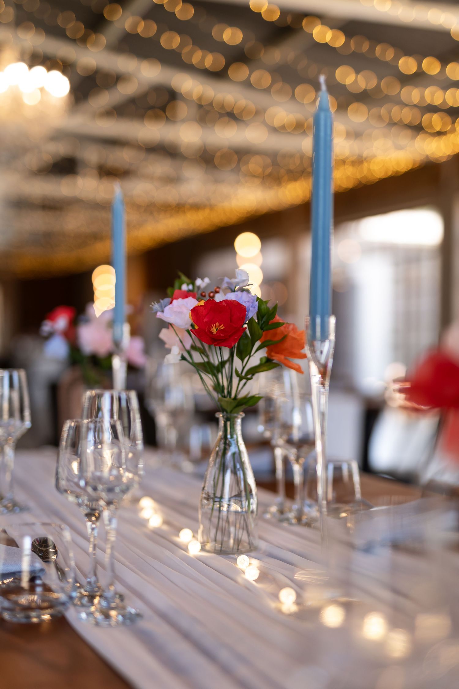 Elegant table setting with blue taper candles and colorful flowers in vases against a backdrop of warm bokeh lights.