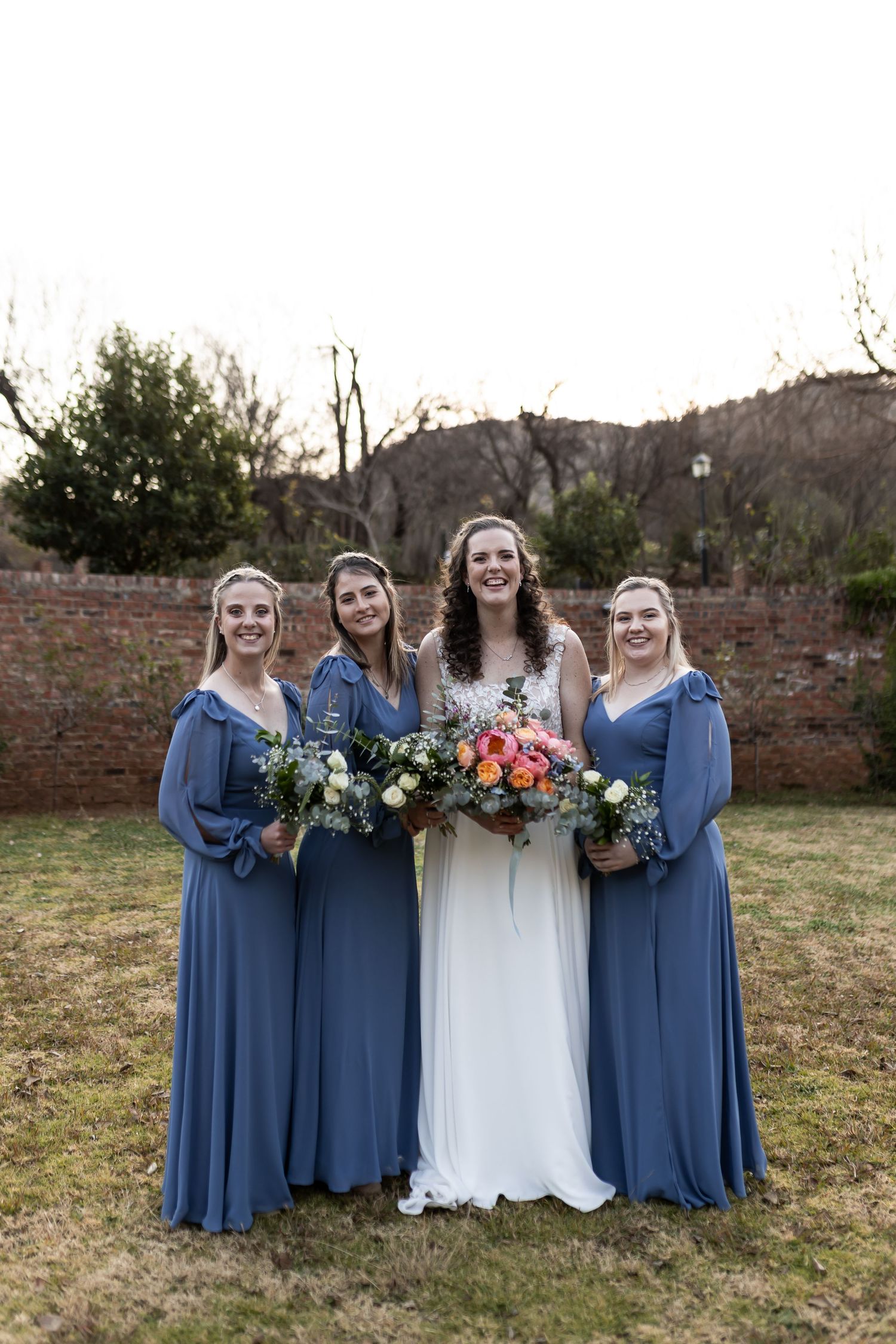 A bride in white stands with bridesmaids wearing blue dresses while holding colorful bouquets in a rustic outdoor setting.
