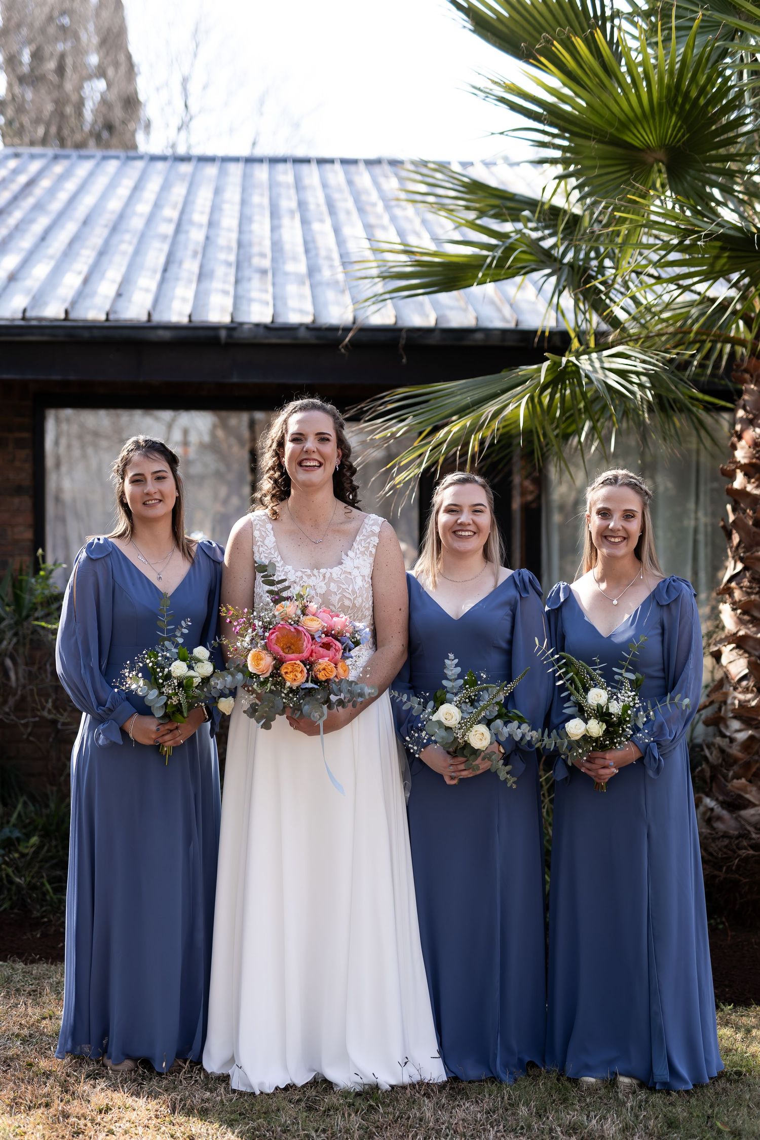 A group stands together outdoors in dusty blue bridesmaid dresses and a white wedding gown holding colorful bouquets.