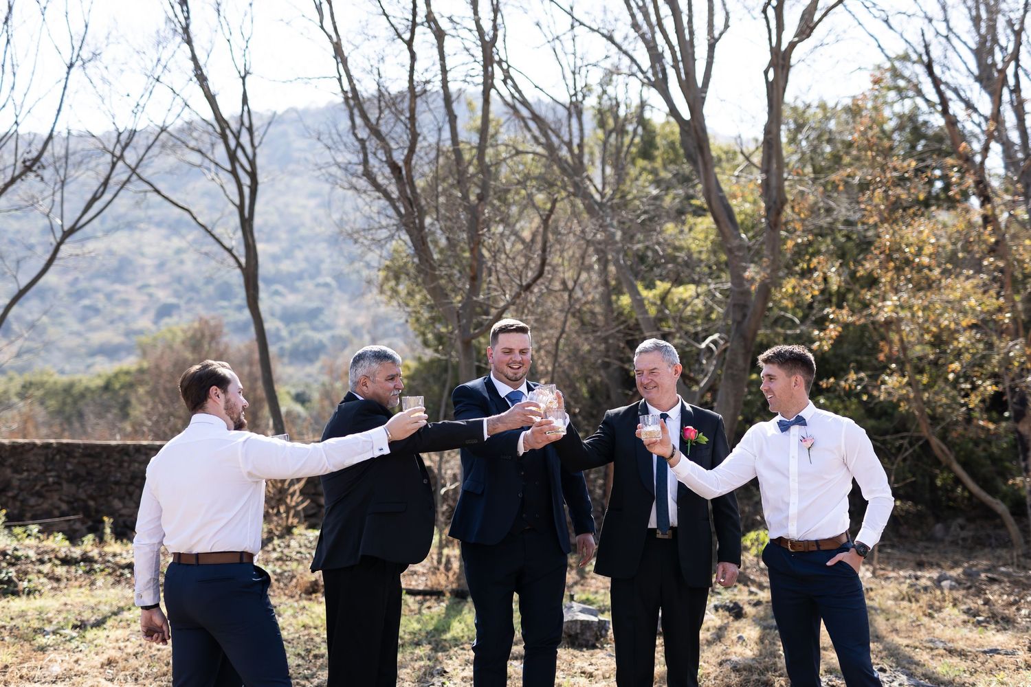 A row of five wedding party members wearing black suits and white shirts pose together against a natural outdoor backdrop.