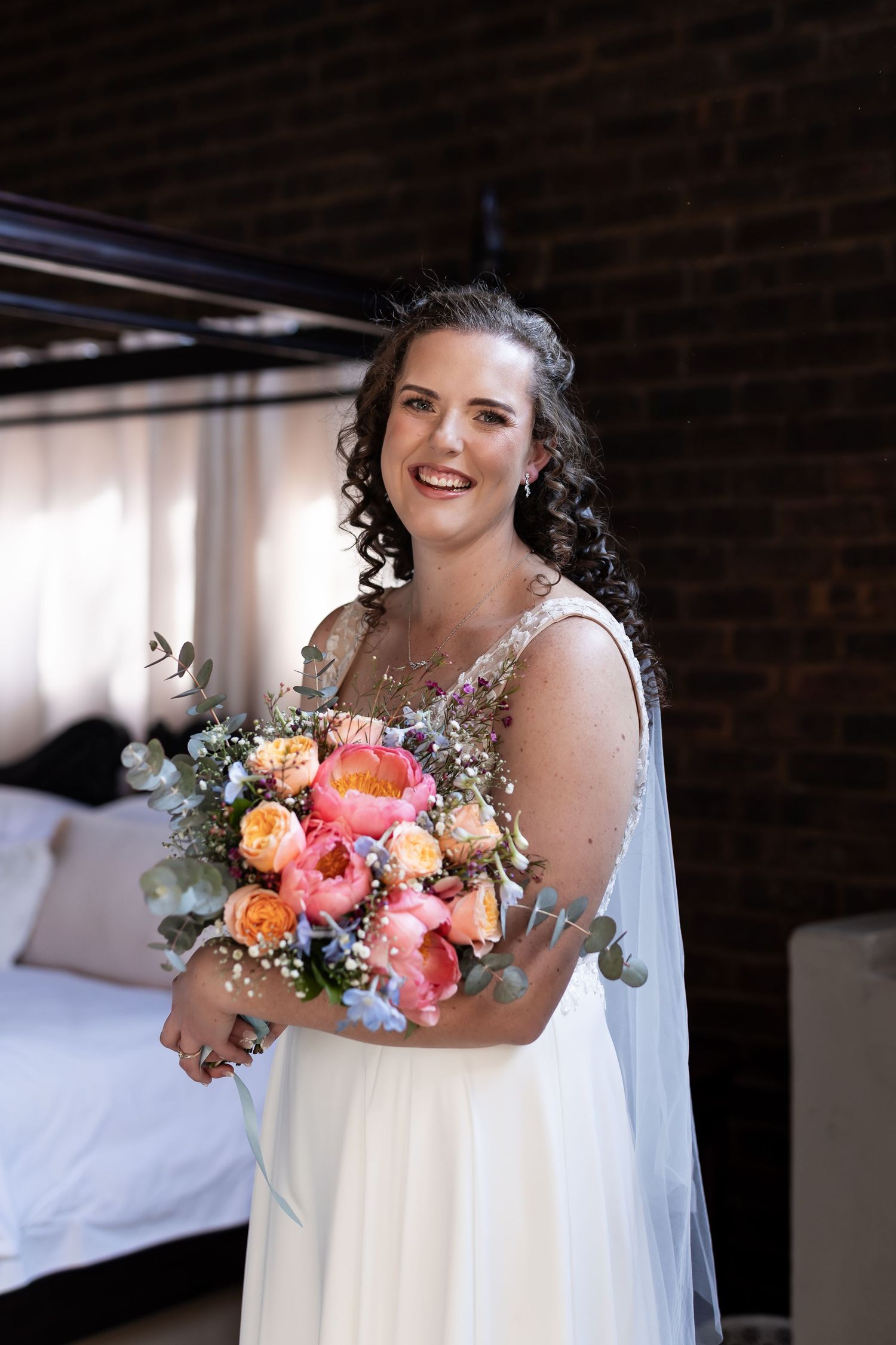 A bride in a flowing white dress holds a vibrant coral and peach wedding bouquet while standing against a brick wall.