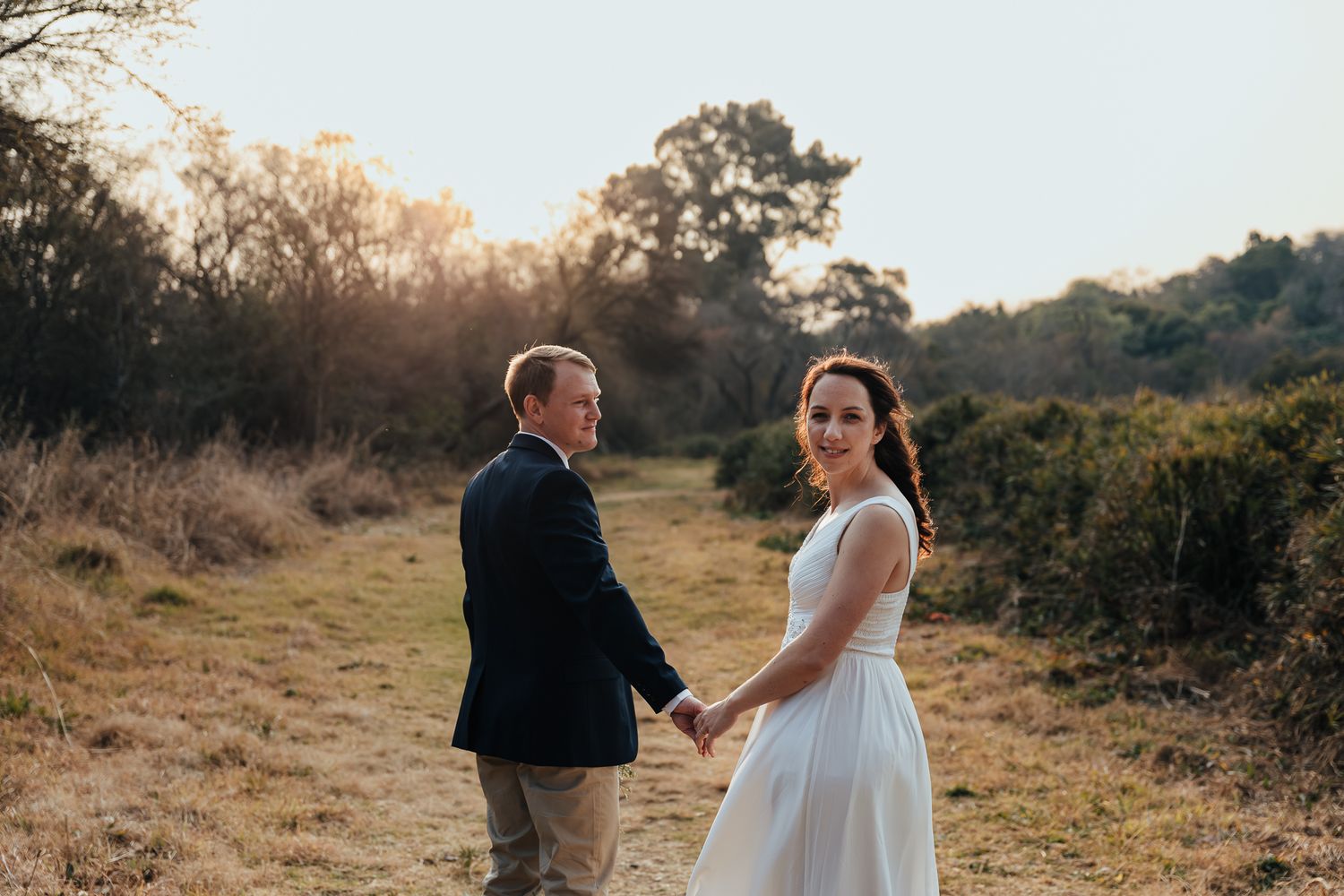 A couple in wedding attire strolling through a golden sunlit field during sunset.