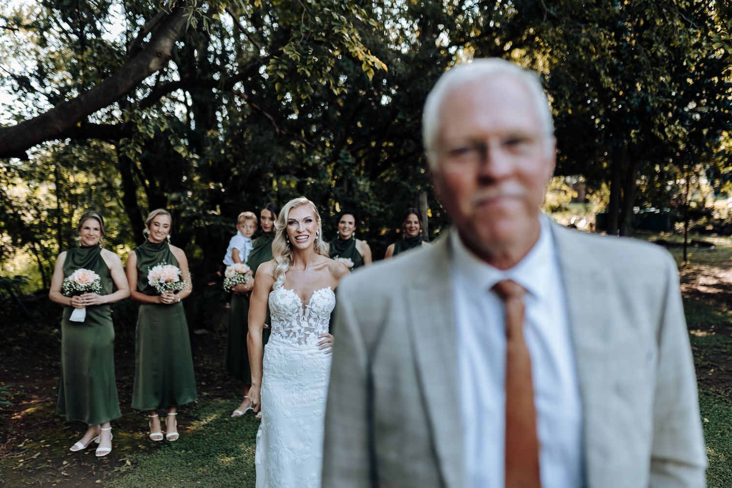 Wedding party in emerald dresses and white gown pose under trees during outdoor ceremony.