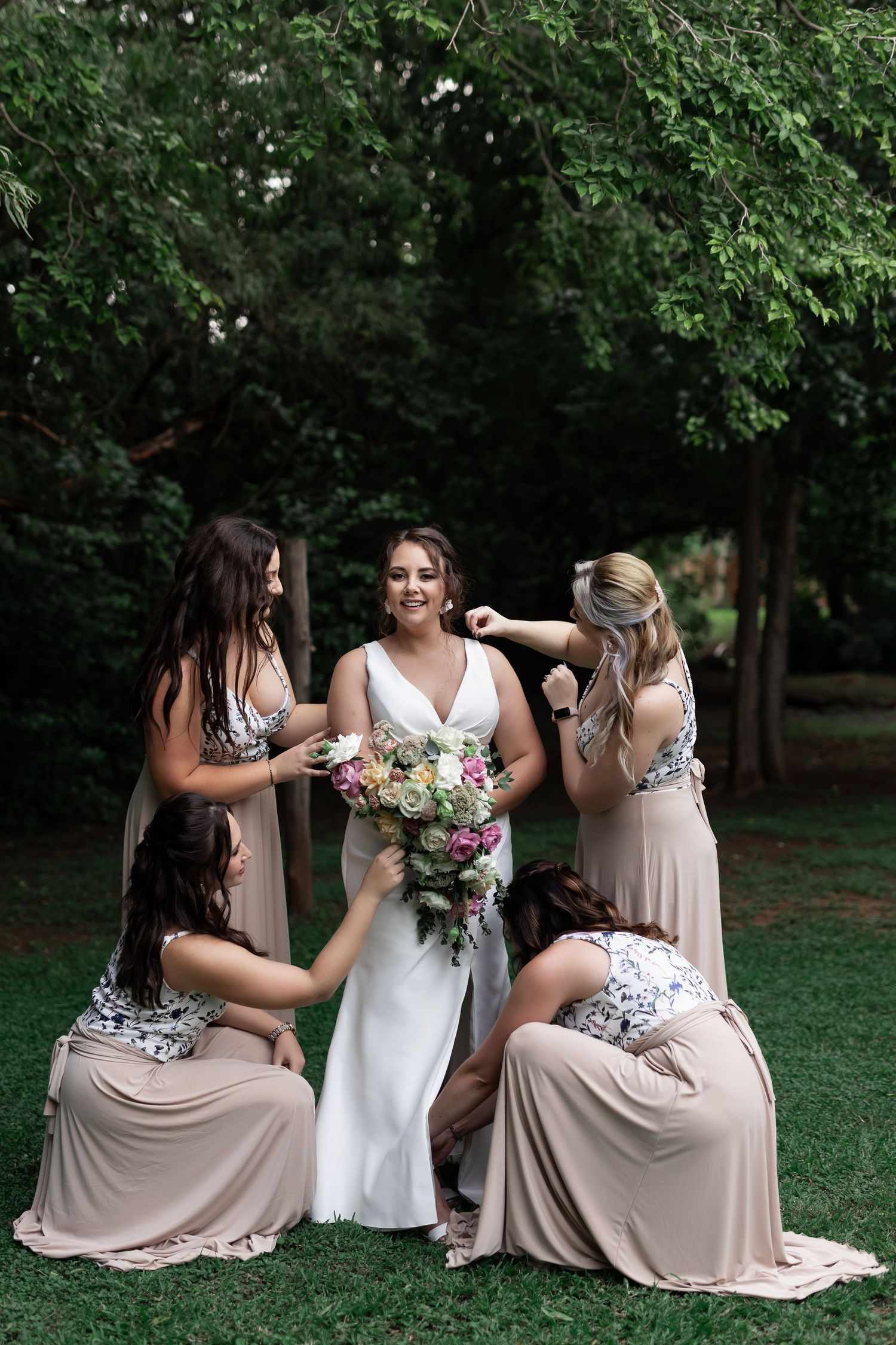 Candid wedding party photo with bridesmaids in blush dresses surrounding bride on grassy lawn.
