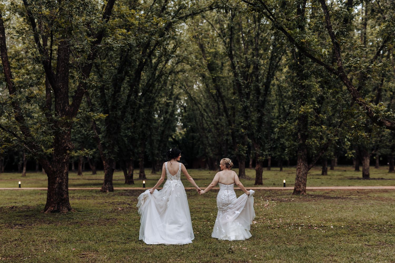 Two people in white wedding dresses embrace among bamboo trees in a garden setting.