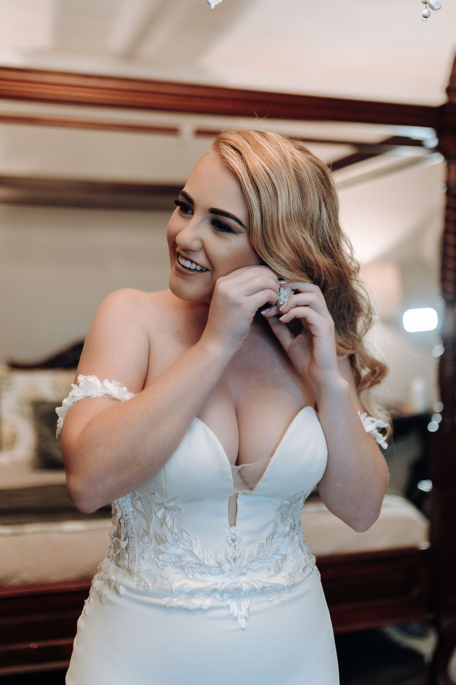 Bride putting on earrings while getting ready in romantic off-shoulder wedding gown.