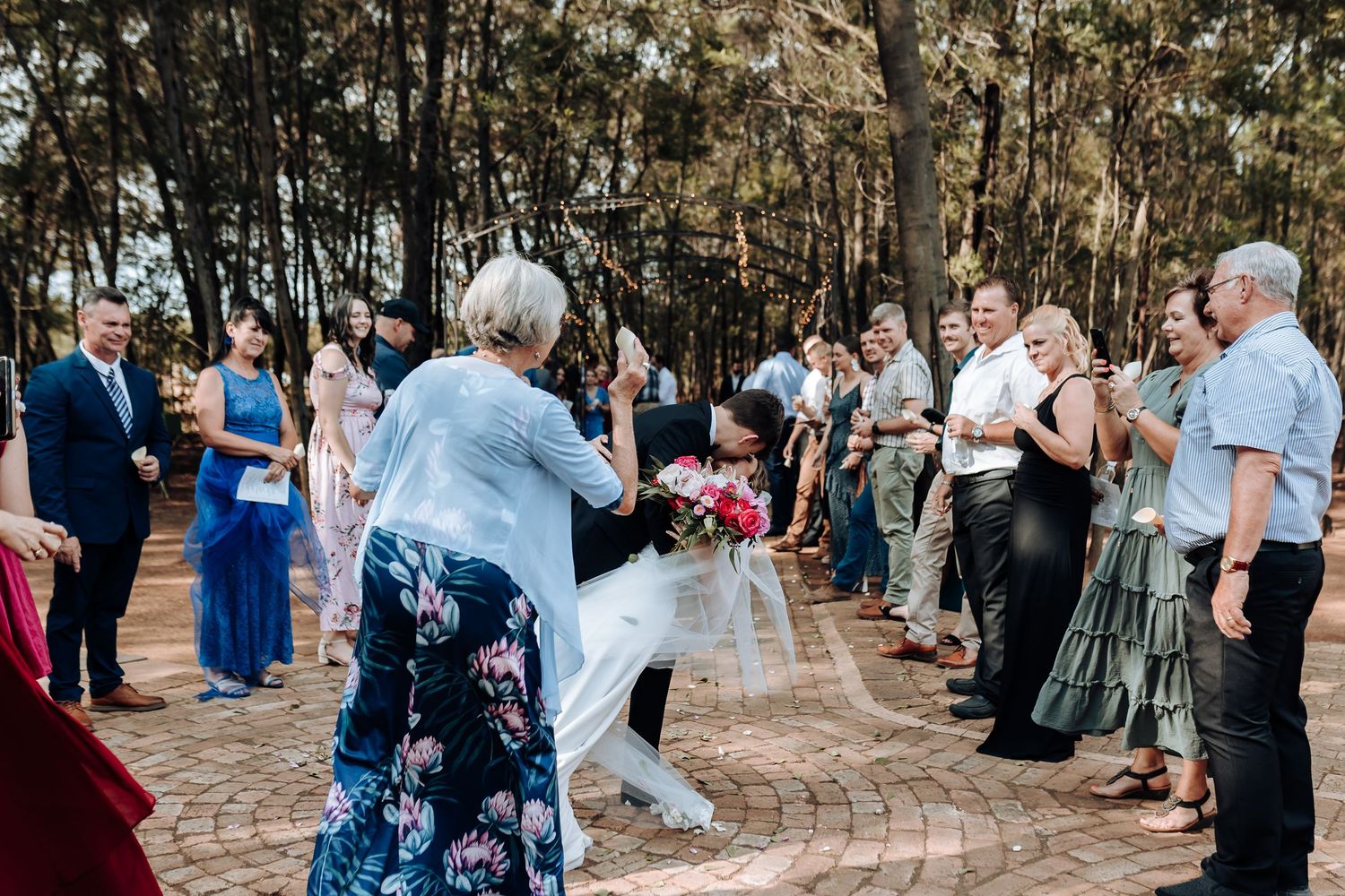 Wedding guests gathered outdoors on brick pathway surrounded by tall trees.