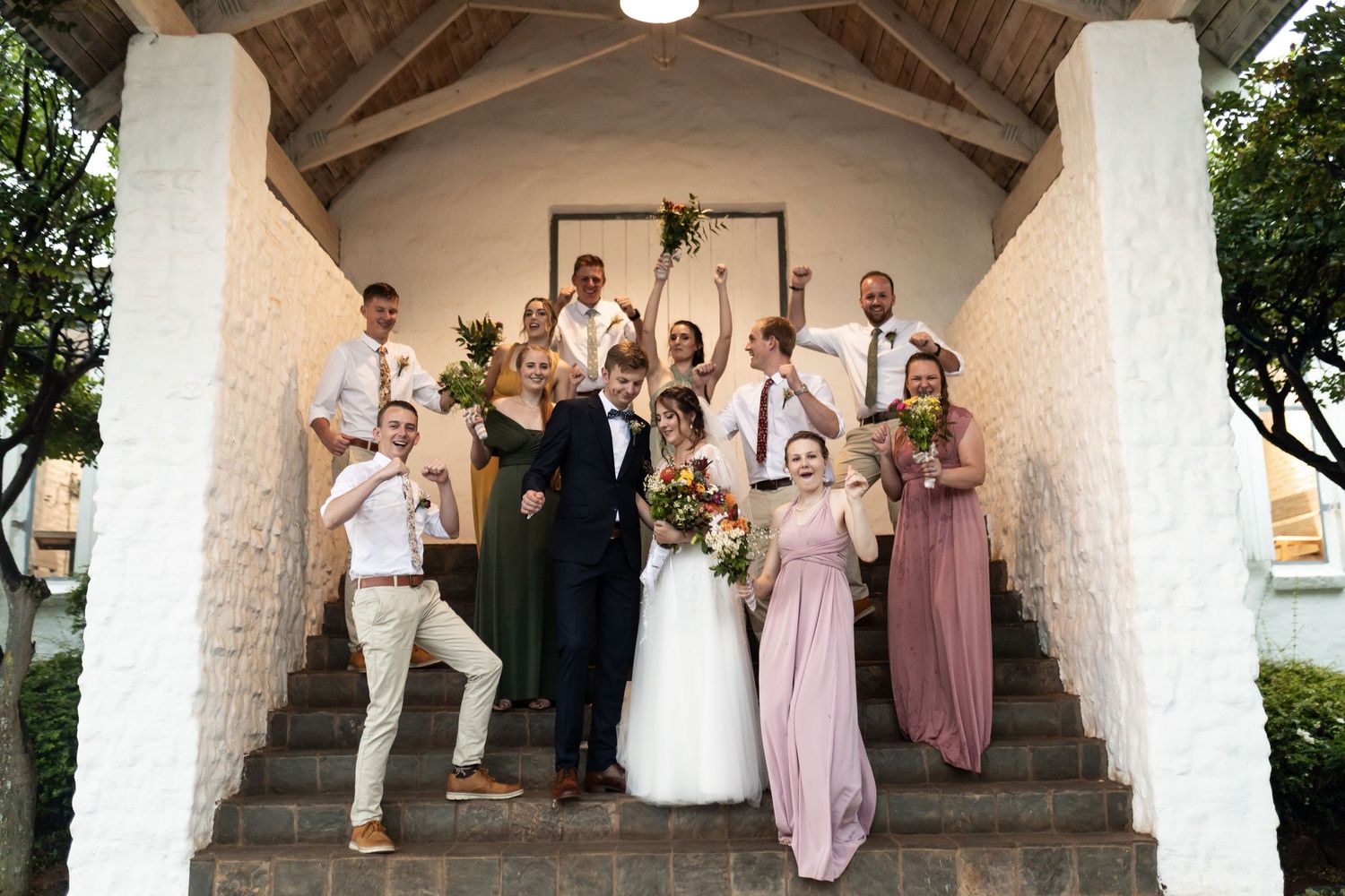 Full wedding party stands on steps of white stone chapel for group photo.