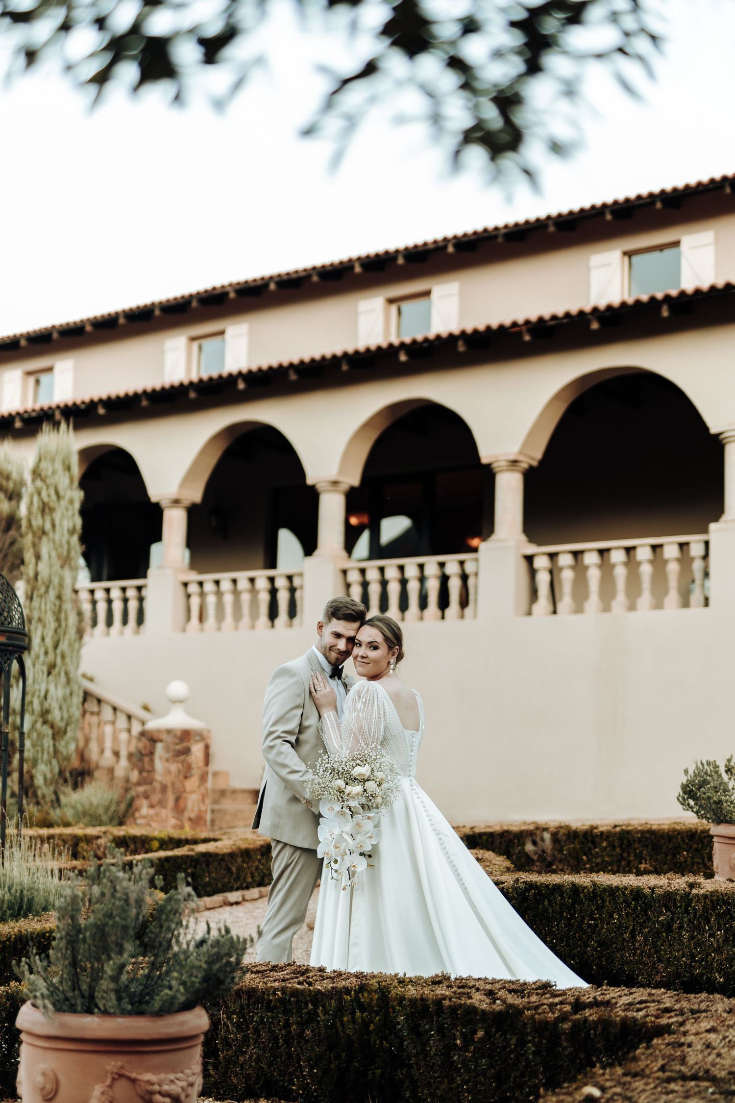 Wedding photograph taken in front of a Mediterranean-style villa with arched walkway.