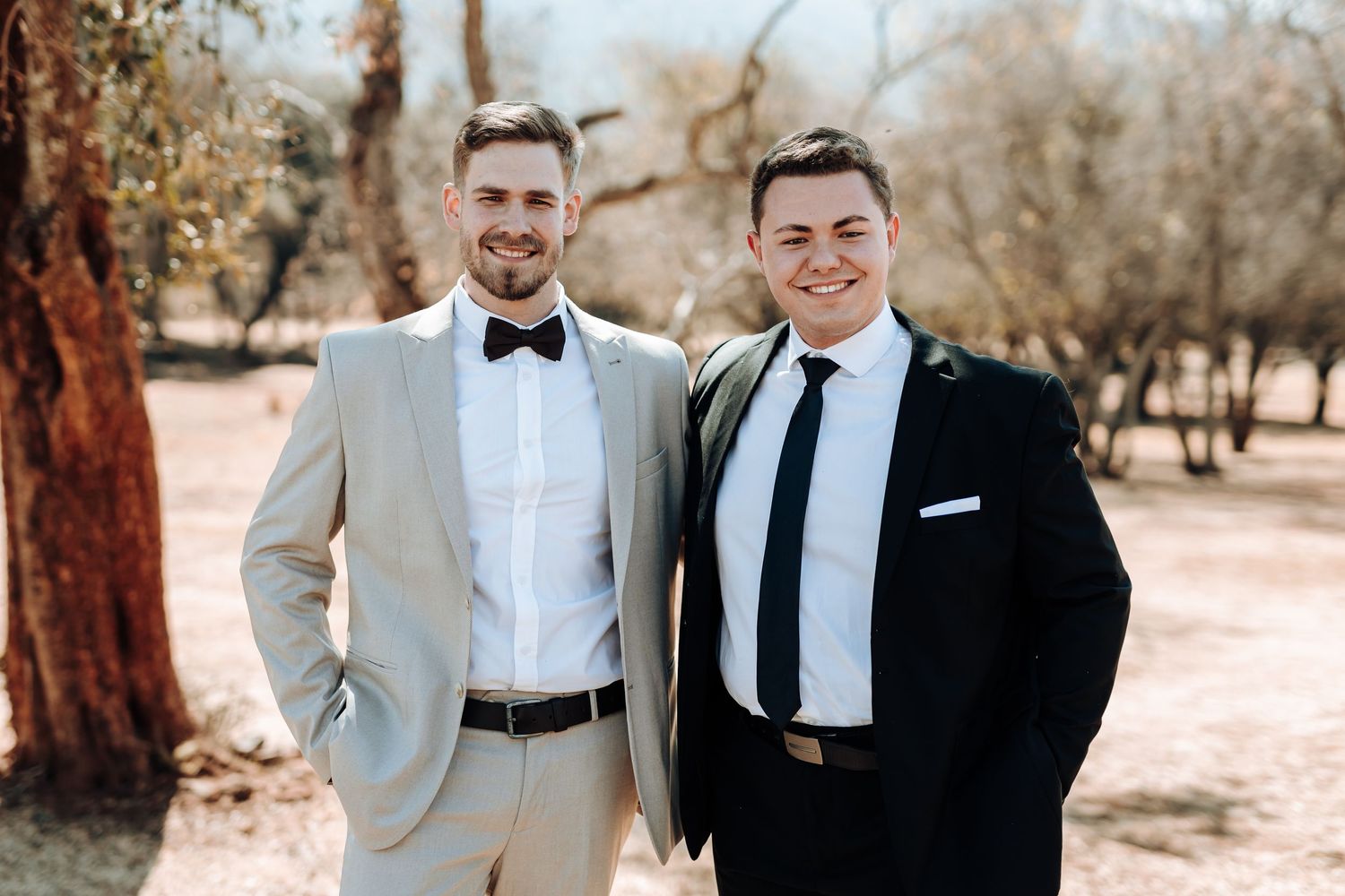Two men in formal attire stand together for a wedding photo in a rustic outdoor setting.