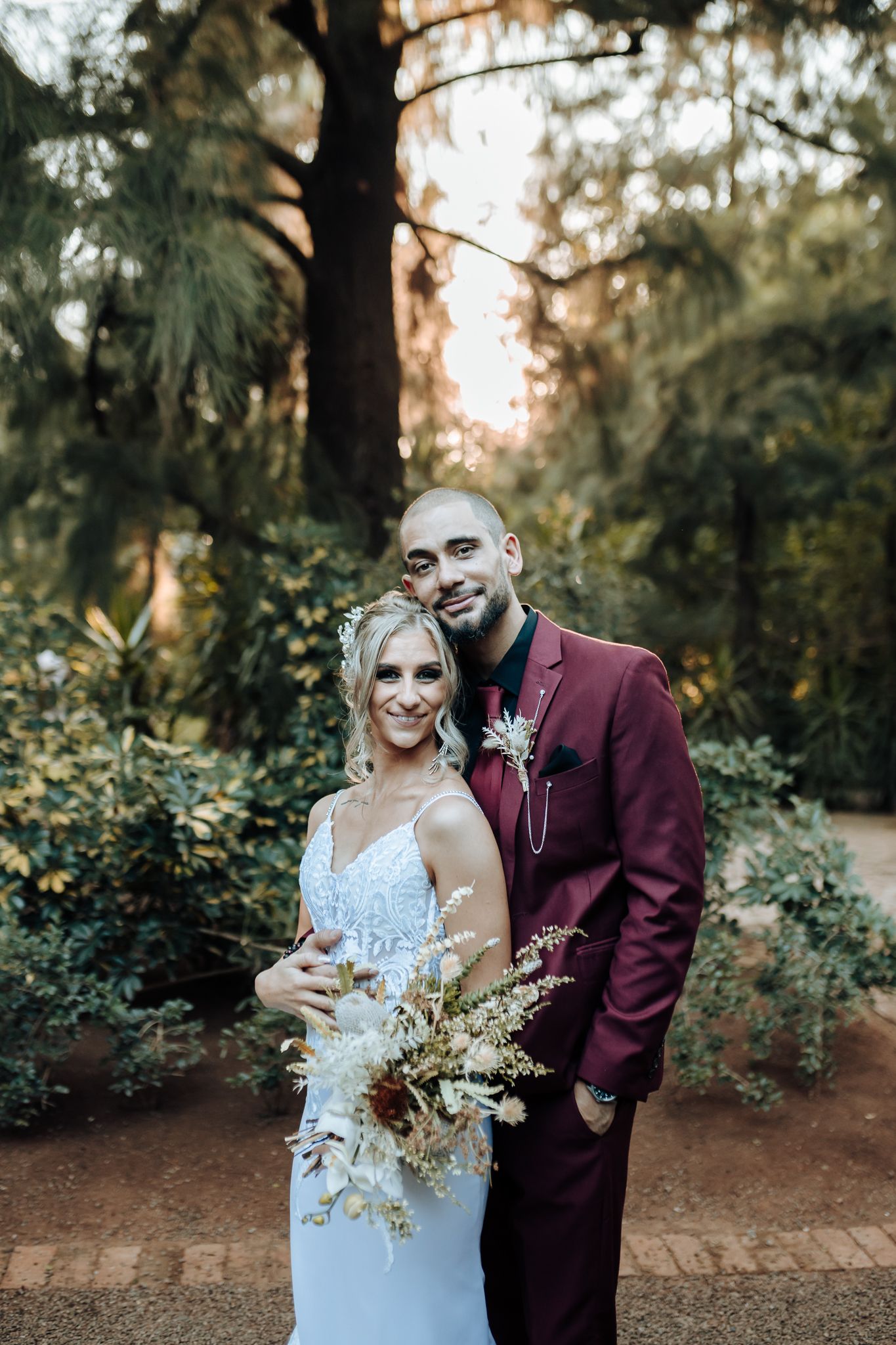 Newlyweds pose together in a garden setting with natural sunlight filtering through trees.
