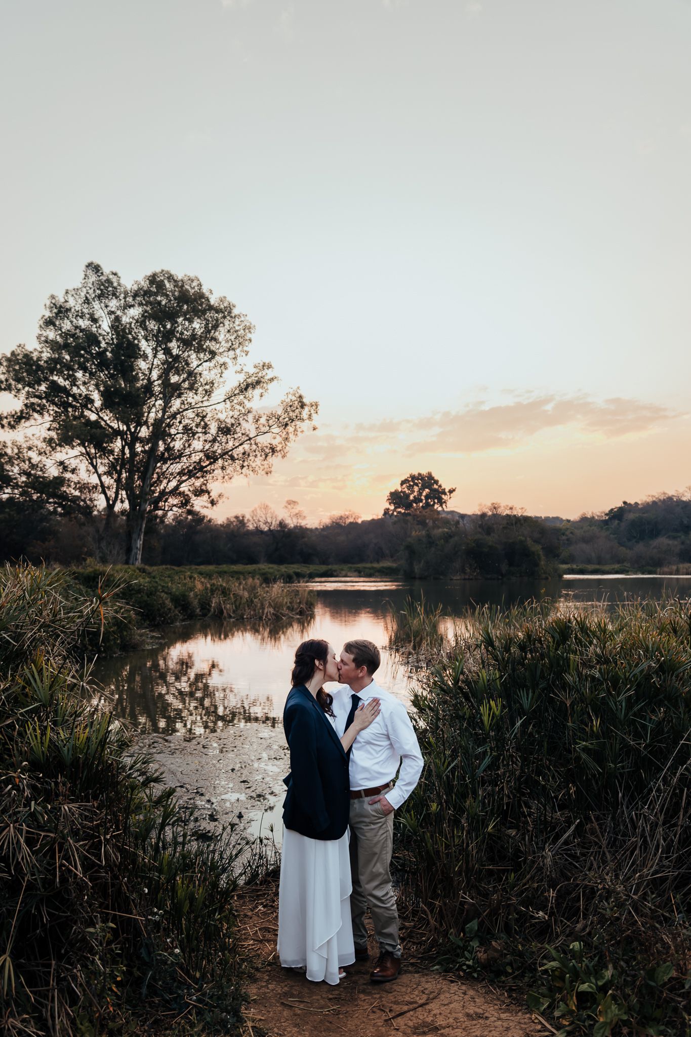 A couple shares an intimate moment by the waterside during golden hour, surrounded by natural landscape.