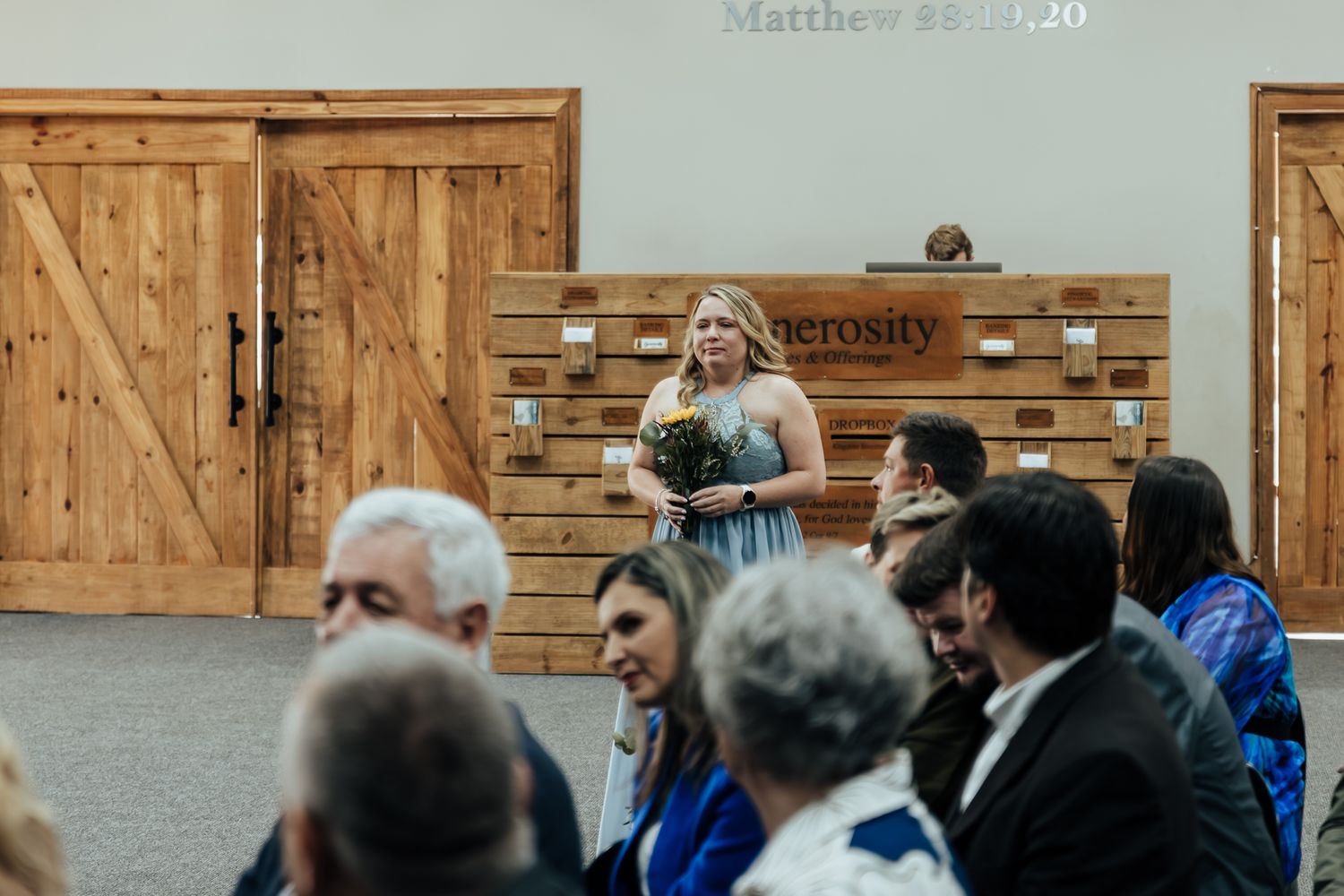 A church service setting with attendees gathered while someone stands on a stage with wooden barn doors in background.
