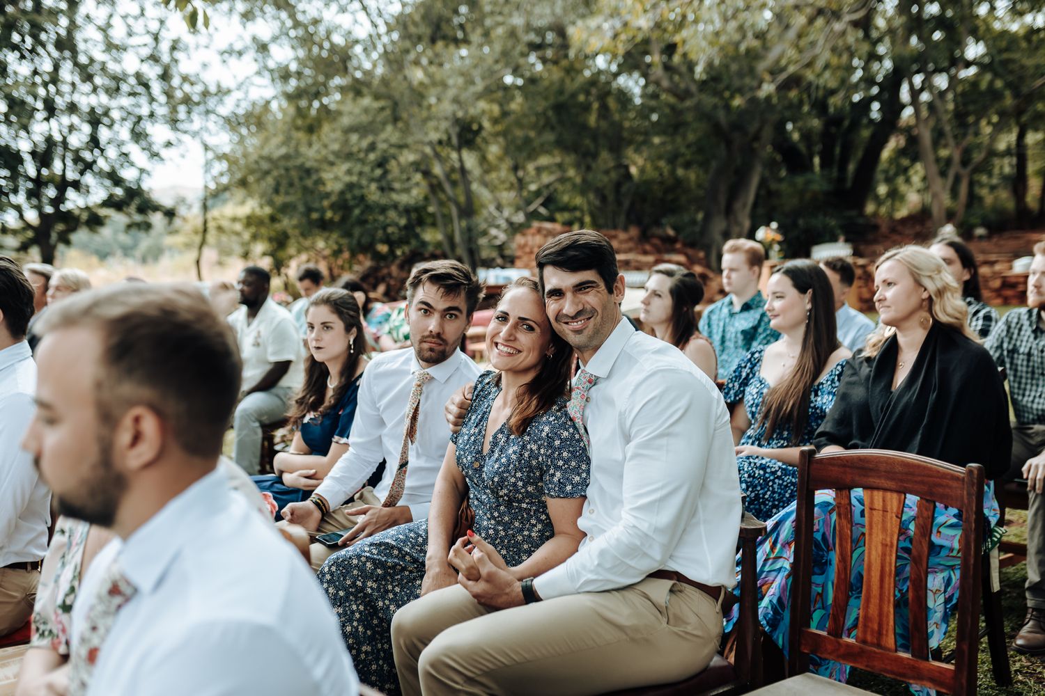 Wedding guests seated on wooden chairs during an outdoor ceremony surrounded by trees.