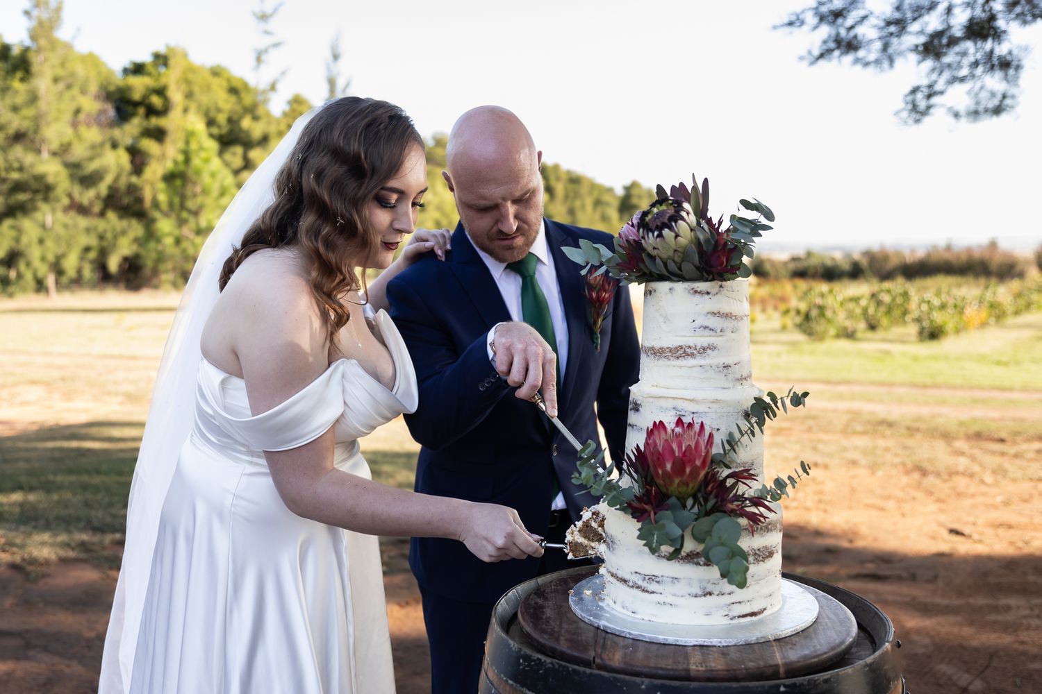 Couple cutting their rustic white wedding cake decorated with native flowers outdoors.