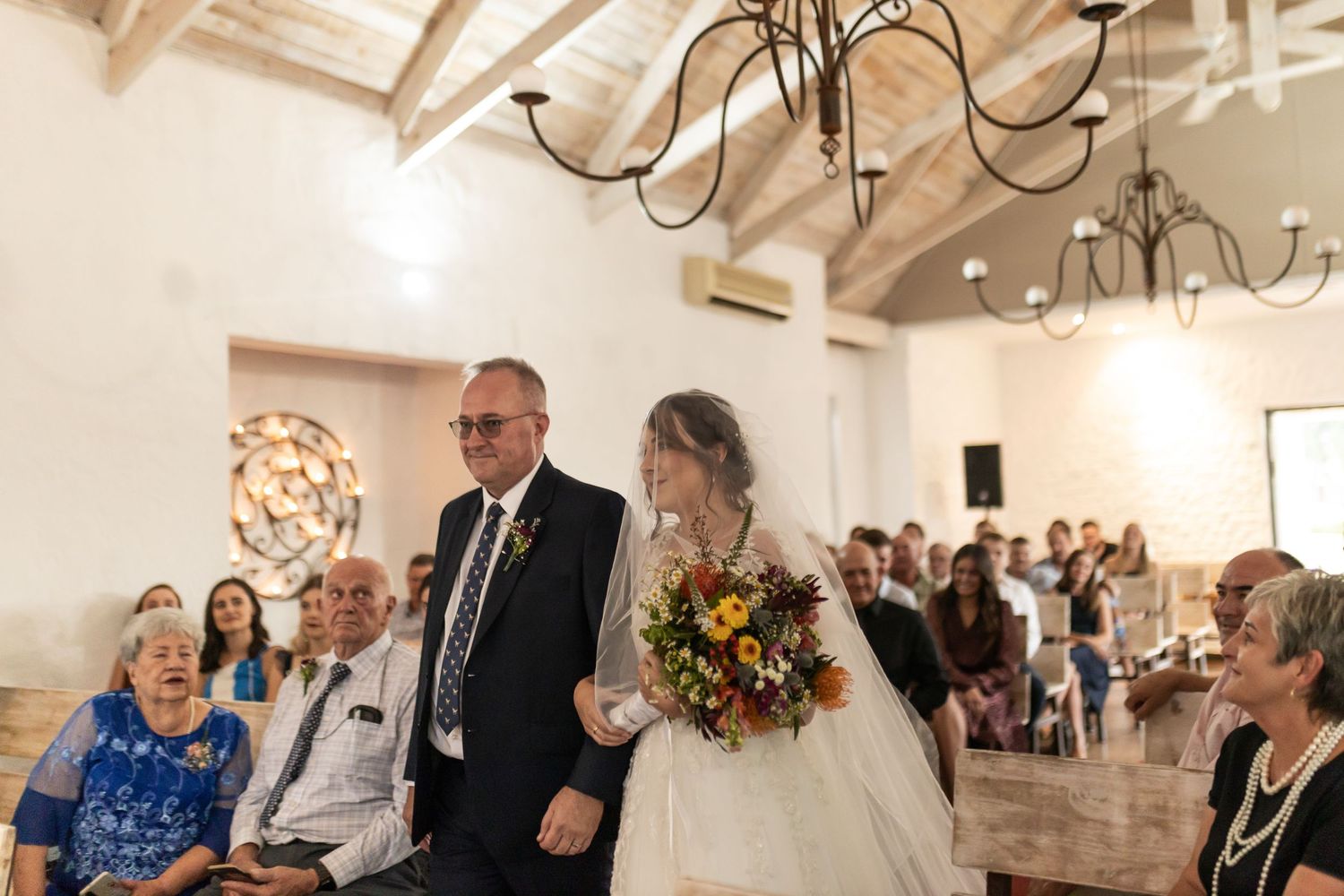 Traditional wedding ceremony procession in chapel with exposed beam ceiling and chandelier lighting.