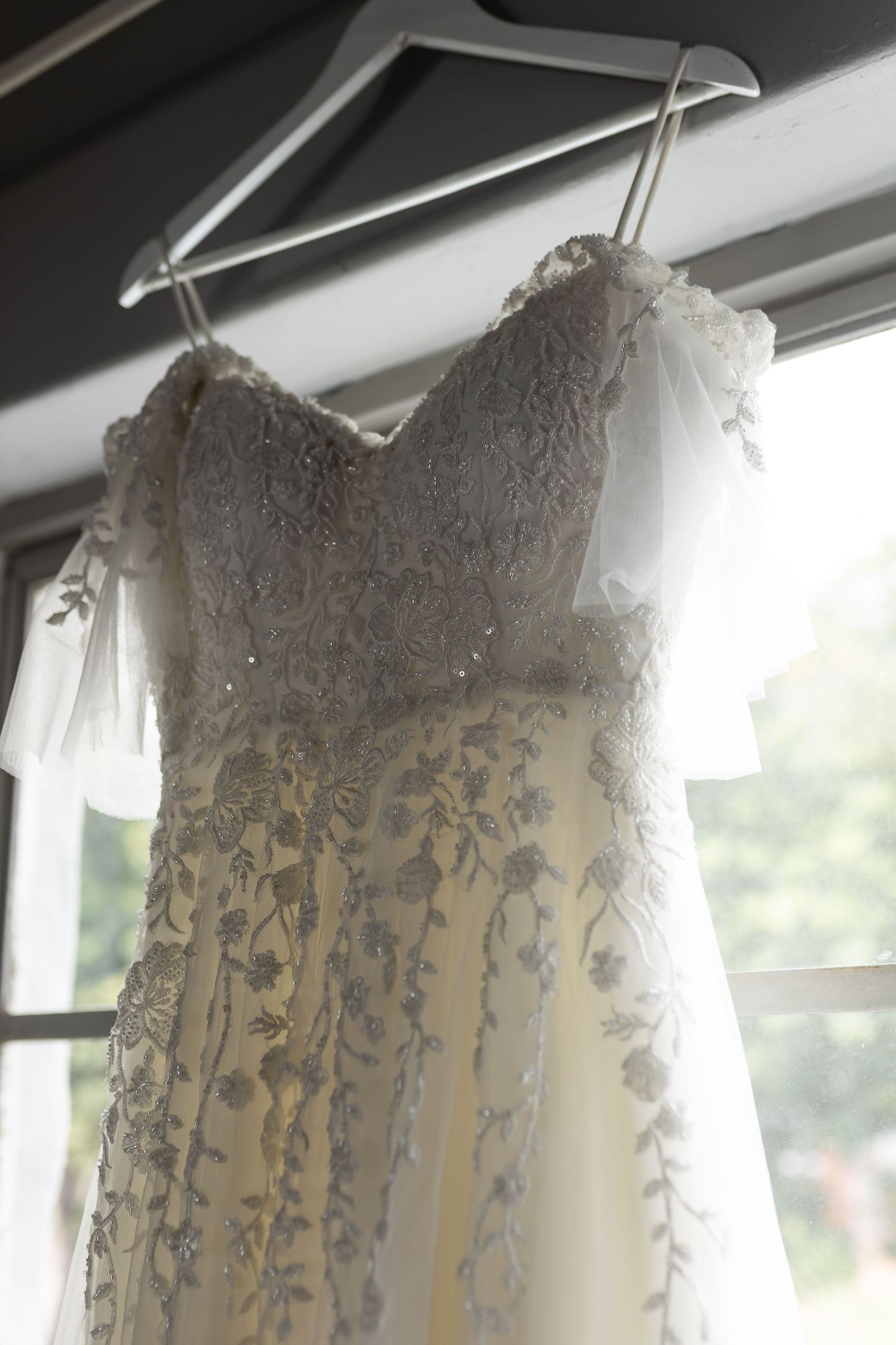 Close-up detail of a white lace wedding dress bodice hanging by a window.
