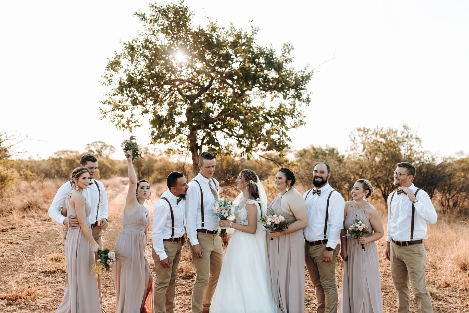 Wedding party in neutral tones poses beneath a tree in rustic outdoor setting at golden hour.