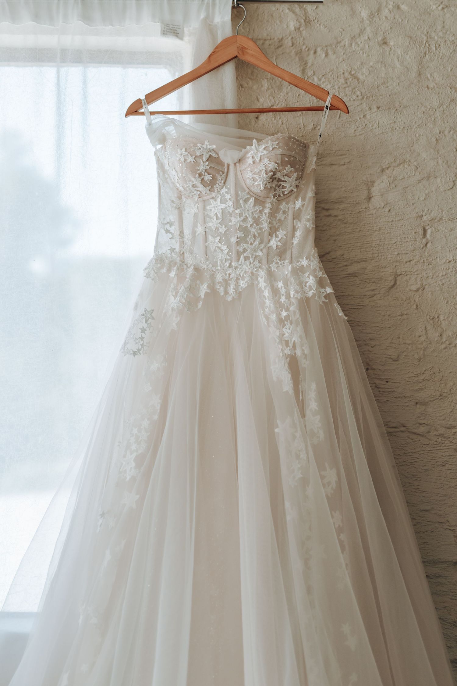 A delicate strapless wedding gown with lace details hangs on a wooden hanger against a white wall.