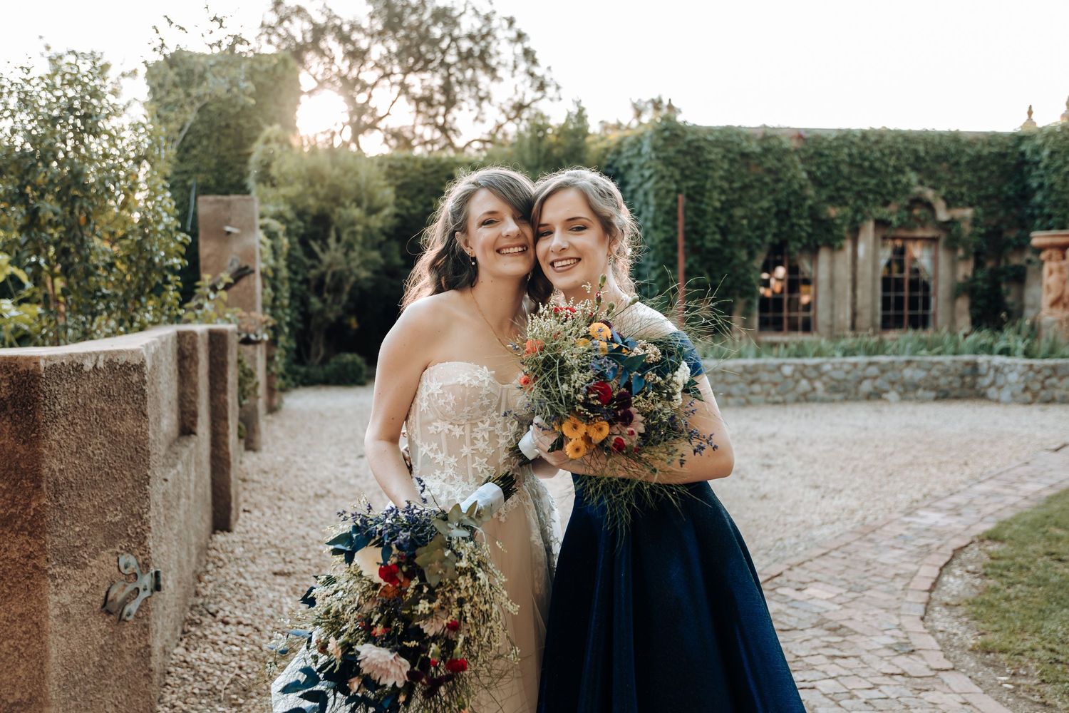 A wedding couple and attendant in navy dress pose together with vibrant florals at sunset in a garden setting.