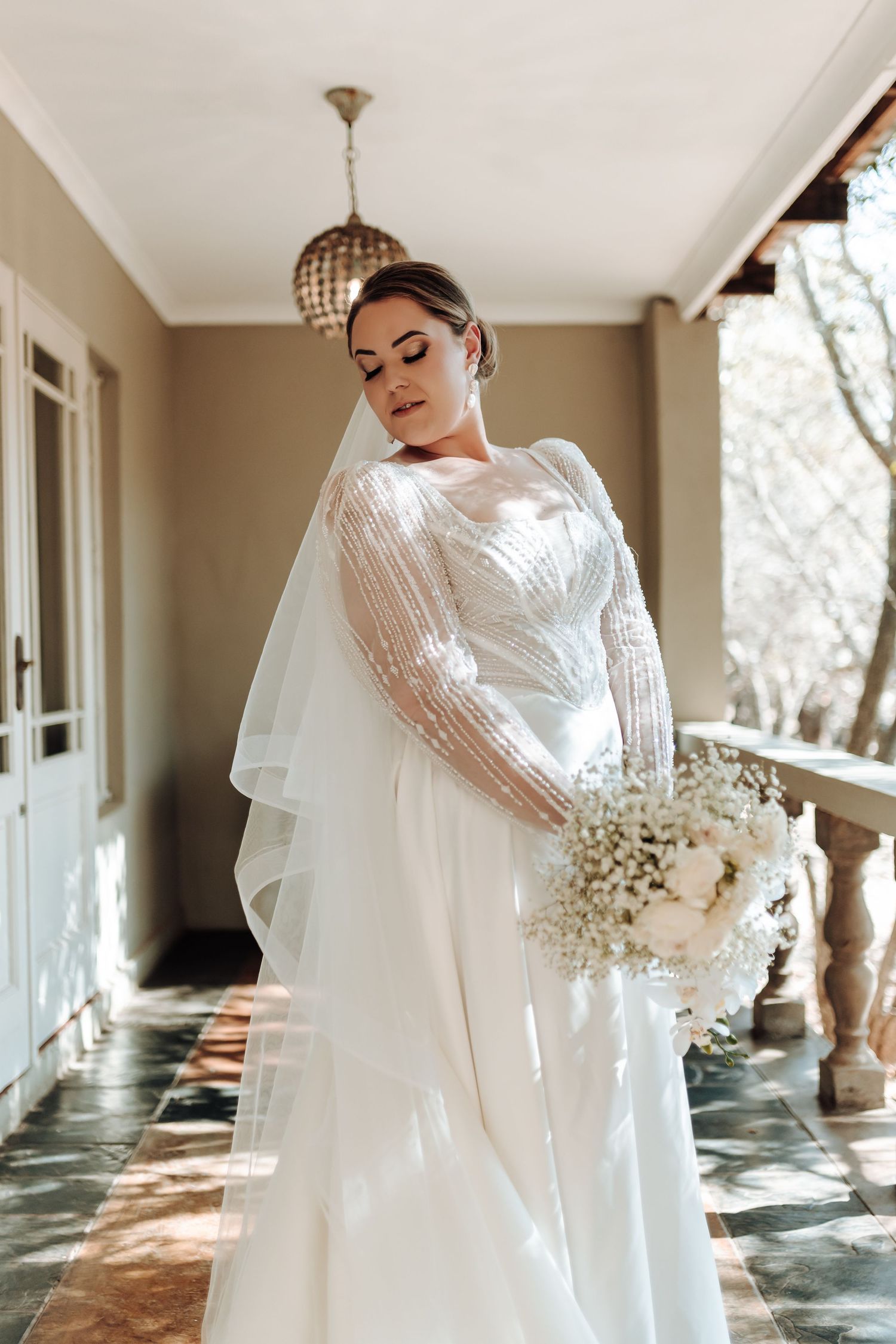 Elegant bride in lace sleeve wedding dress holds baby's breath bouquet on classic porch.