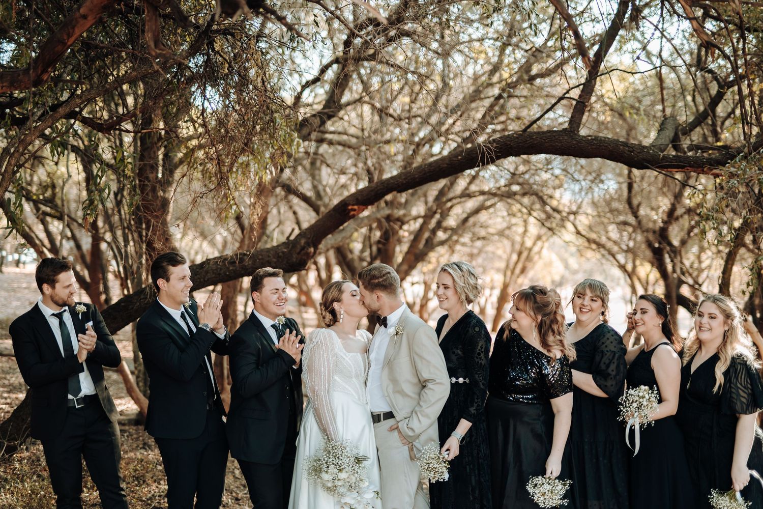 Wedding ceremony under trees with wedding party in black attire gathered around couple.