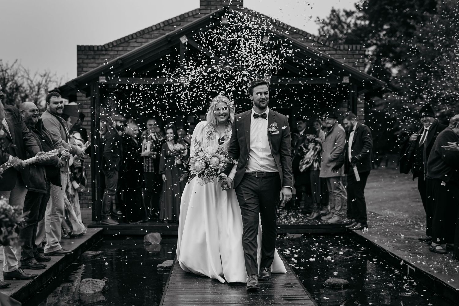 Wedding couple walks together while confetti rains down outside a rustic venue in black and white.