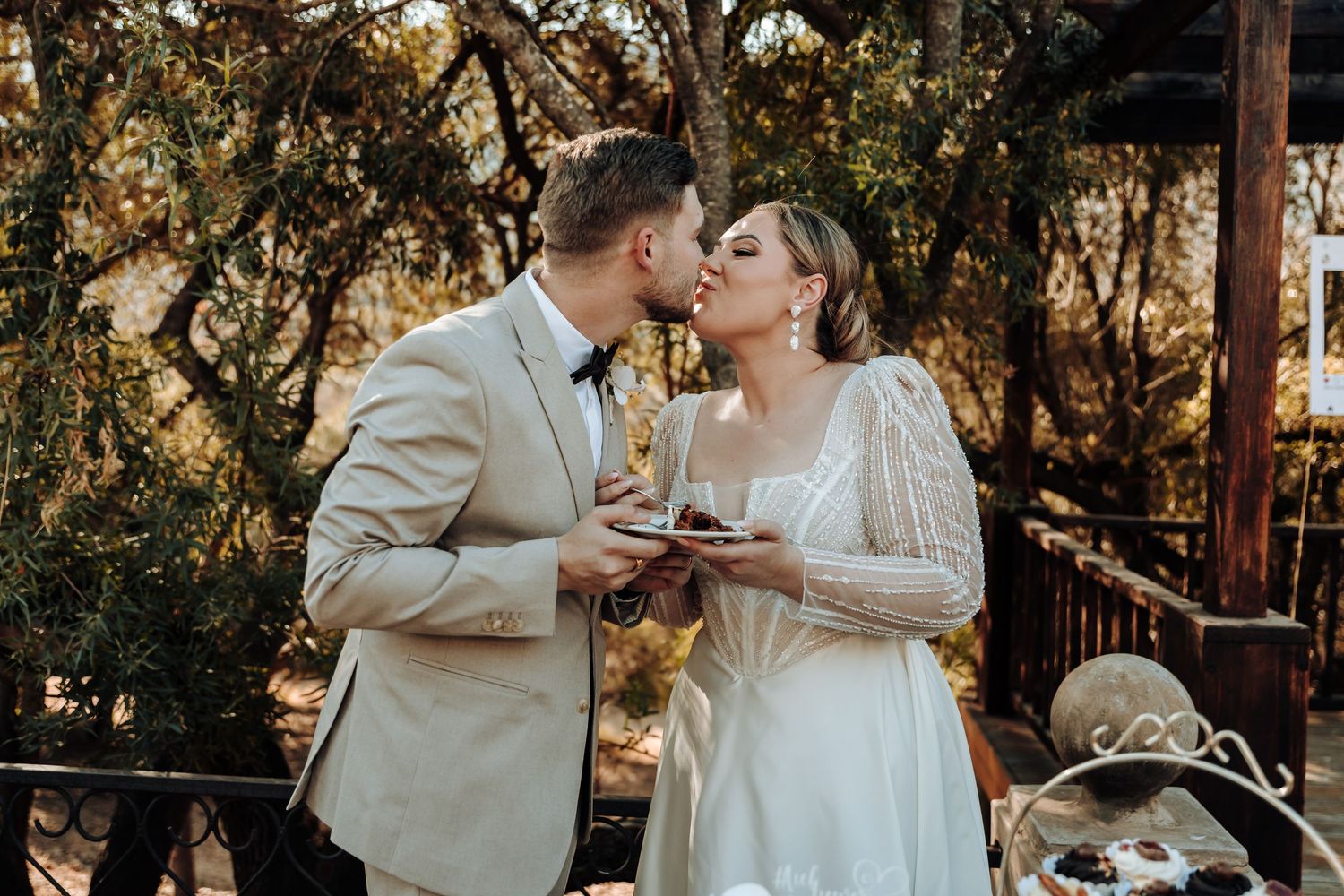 A couple samples wedding cake together at their outdoor reception under trees at golden hour.