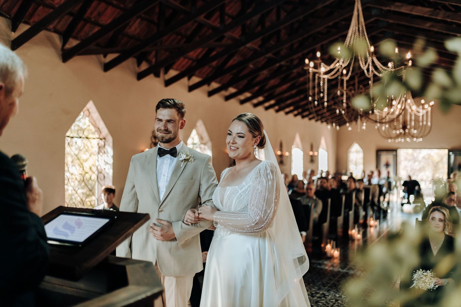 A wedding couple stands at the altar during their ceremony in a beautifully lit chapel with exposed beams.