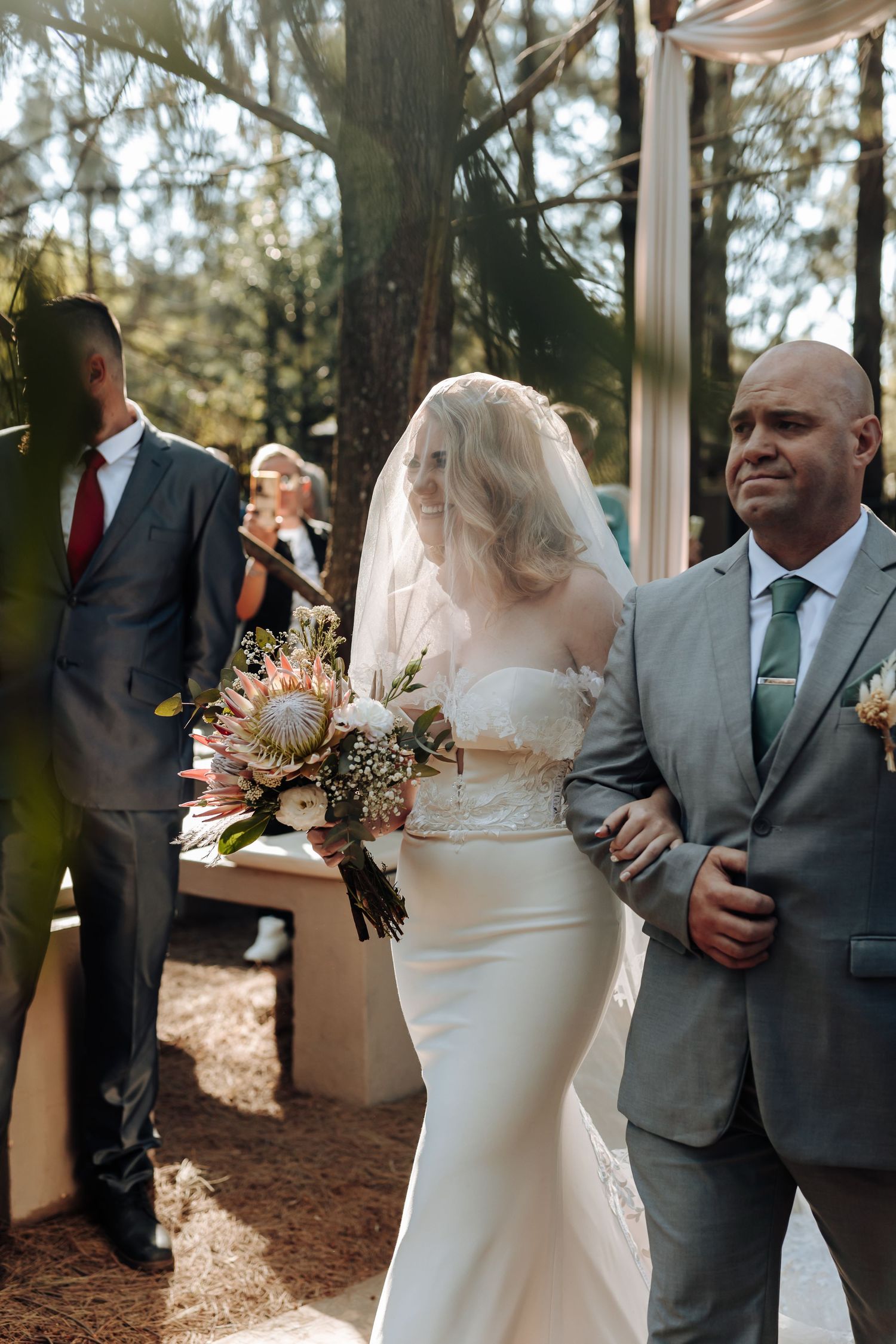 A bride holding a bouquet walks down the aisle during an outdoor forest wedding ceremony.