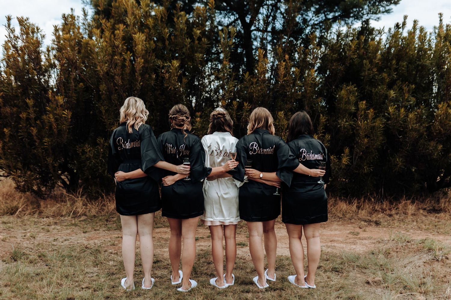 Bridal party shows off matching black robes with 'bridesmaid' text in an outdoor setting.