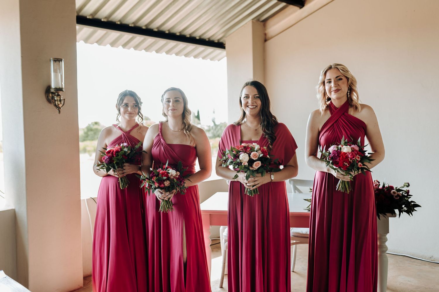 Bridesmaids in matching red formal gowns hold bouquets at outdoor wedding venue.
