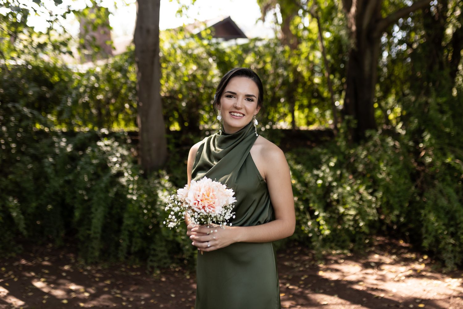A person in a green halter dress poses with a pink floral bouquet in a garden setting.