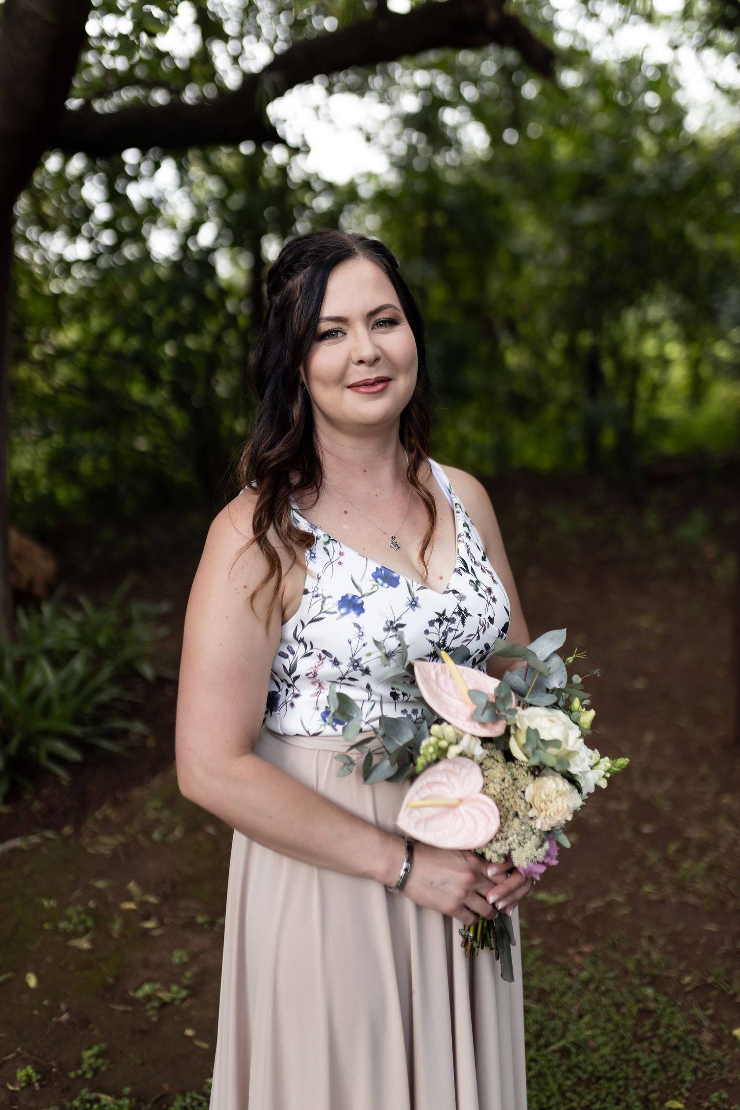 Bridesmaid poses with pastel bouquet wearing floral top and neutral skirt in garden setting.