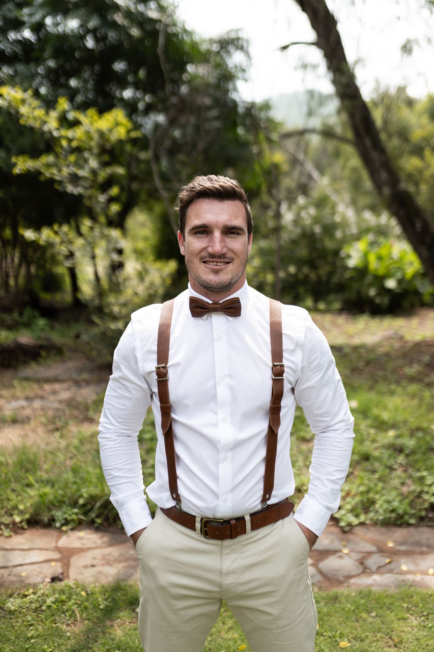 Groomsman in classic wedding attire with brown suspenders and bow tie poses in garden setting.
