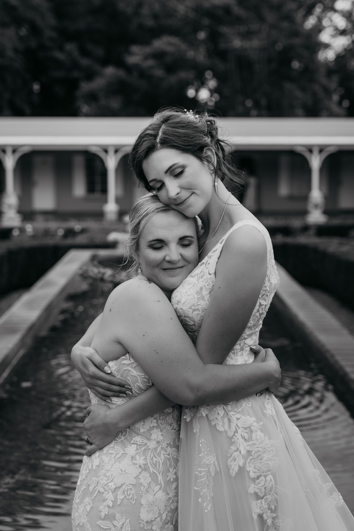 Two people in white lace wedding dresses share a tender embrace in a garden setting.
