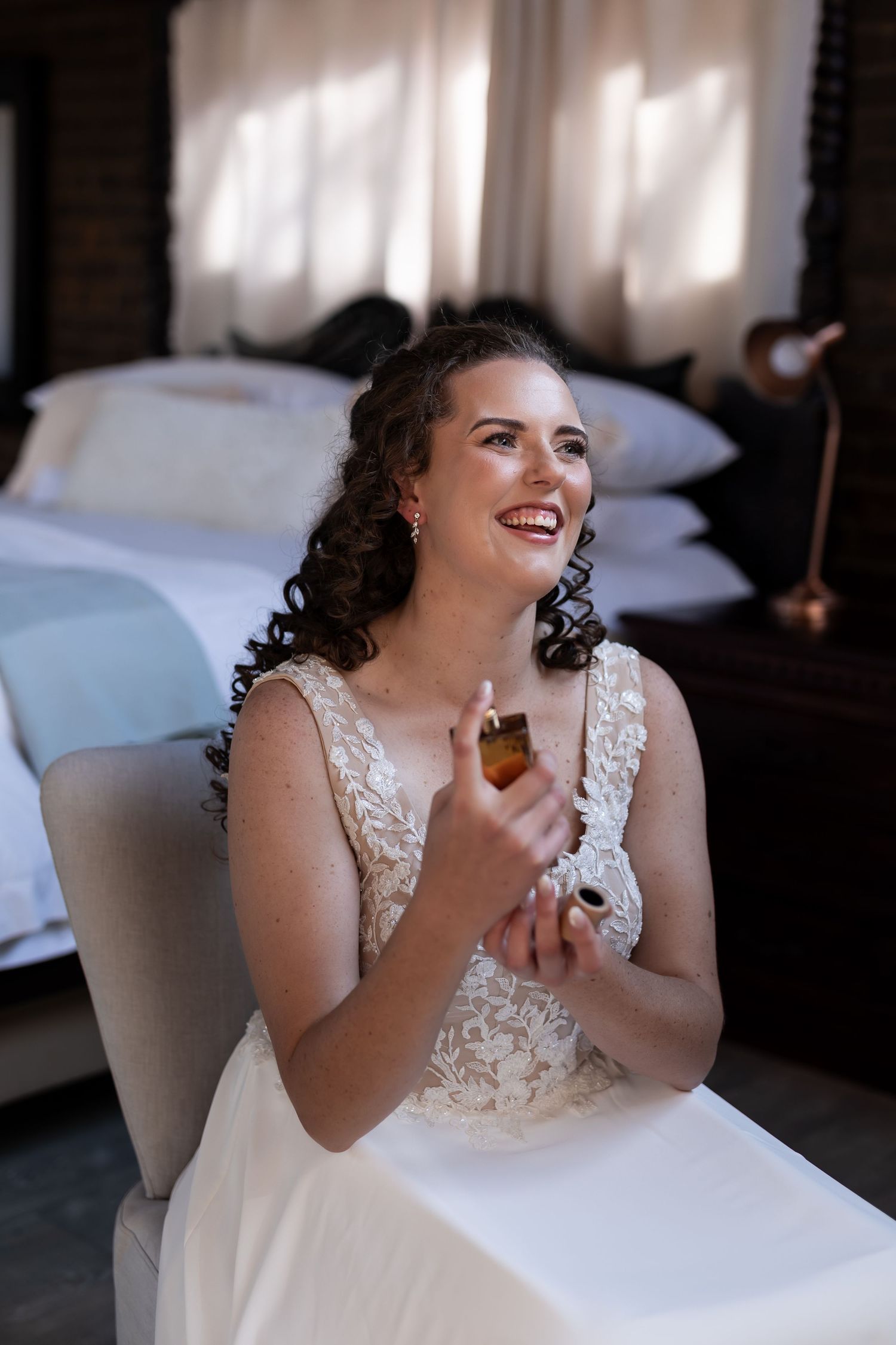 A bride in a lace wedding dress laughs joyfully while seated in an elegant white chair.