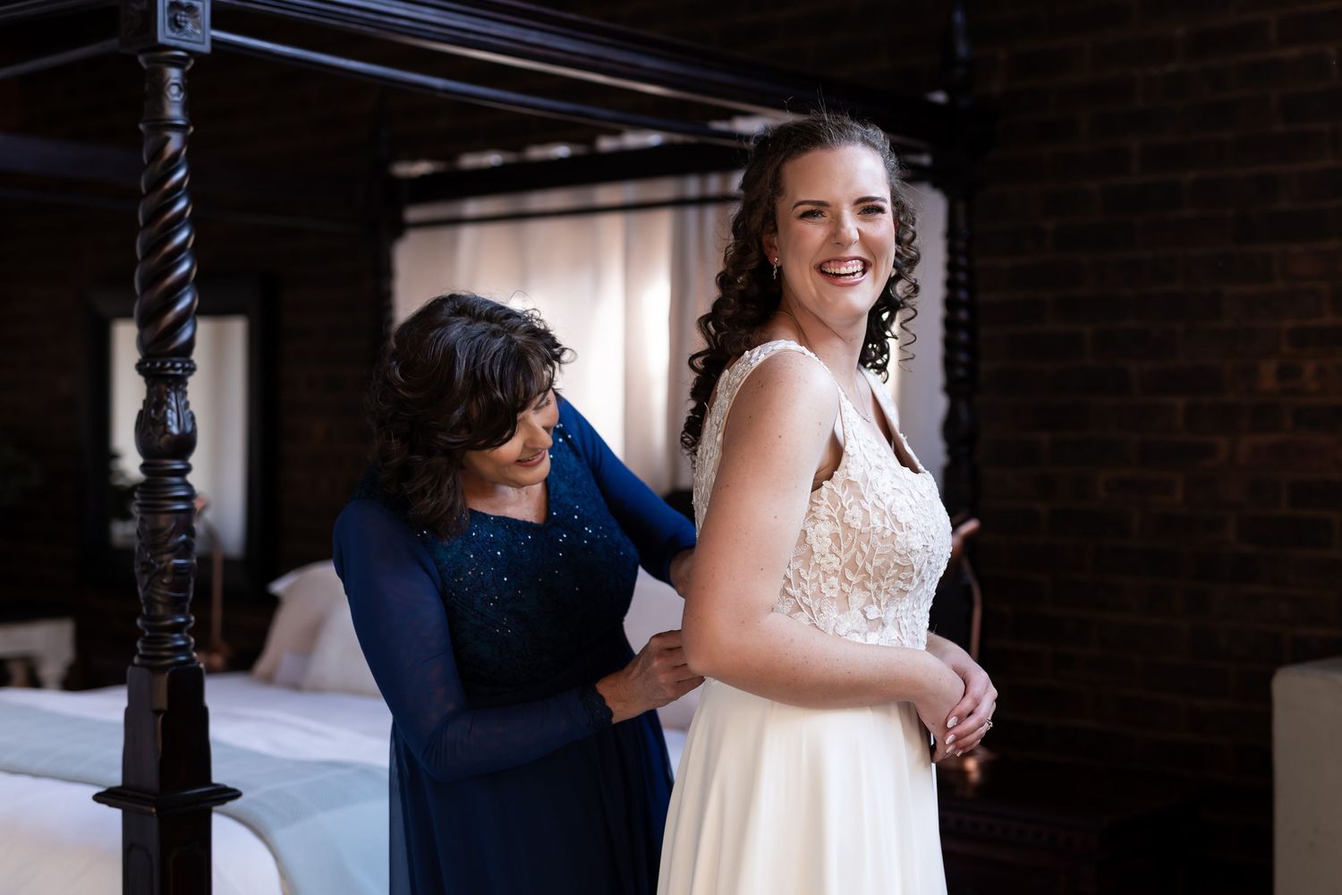 A candid moment during wedding preparations shows two women laughing together, one in a white gown and one in navy blue.