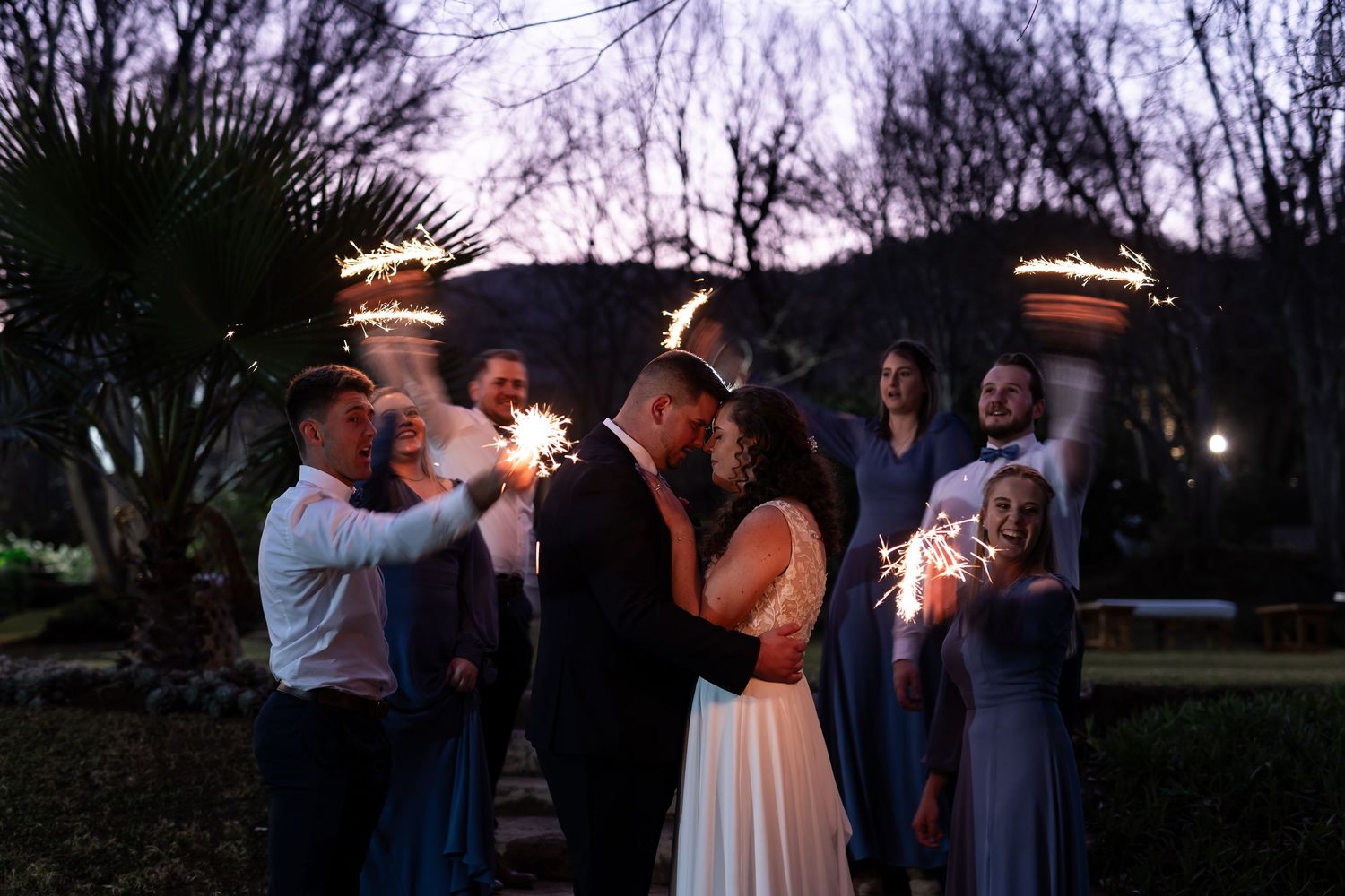 Wedding guests hold sparklers in a circle during a nighttime outdoor celebration, creating a magical lighting effect.