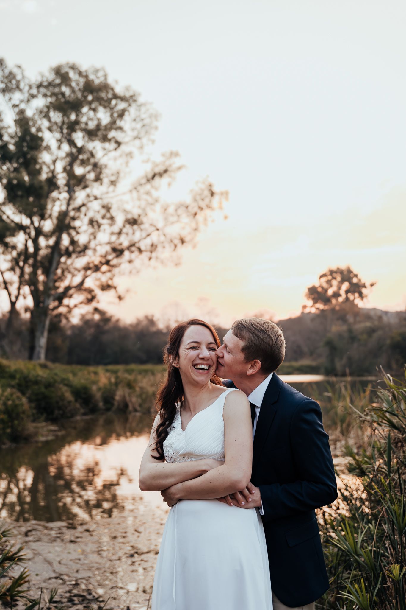 A wedding couple shares a tender moment by a pond during golden hour with a sunset backdrop.