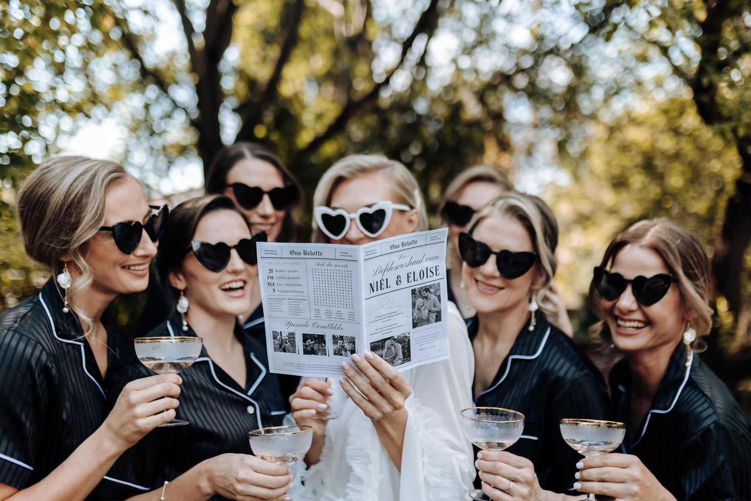 Group wearing sunglasses reads wedding newsletter while holding champagne glasses outdoors.