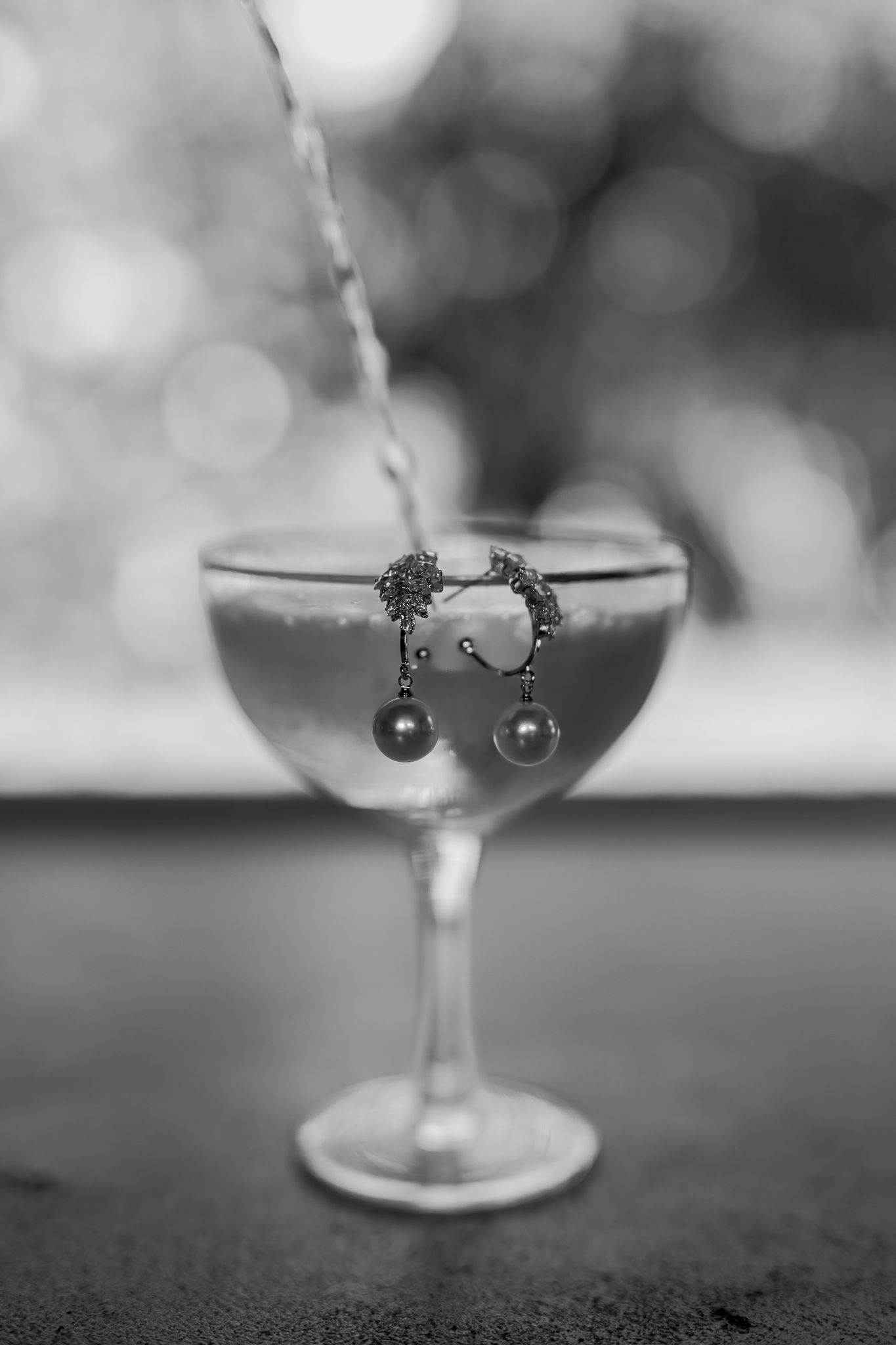 Earrings suspended over champagne glass with bokeh background in black and white.