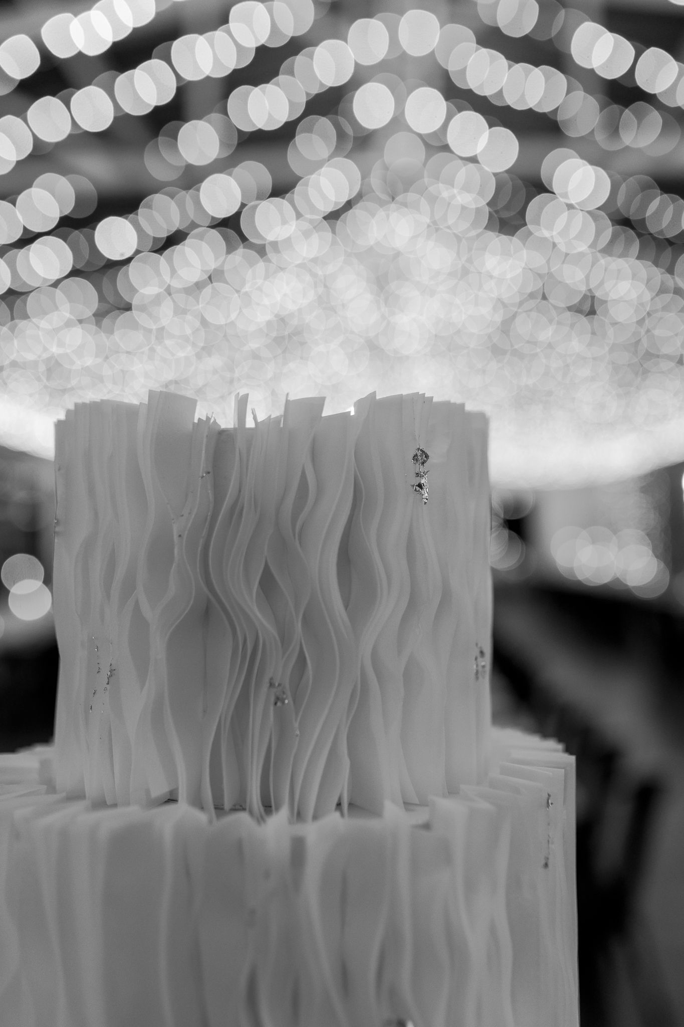 White textured wedding cake with wavy fondant design captured against bokeh lights.