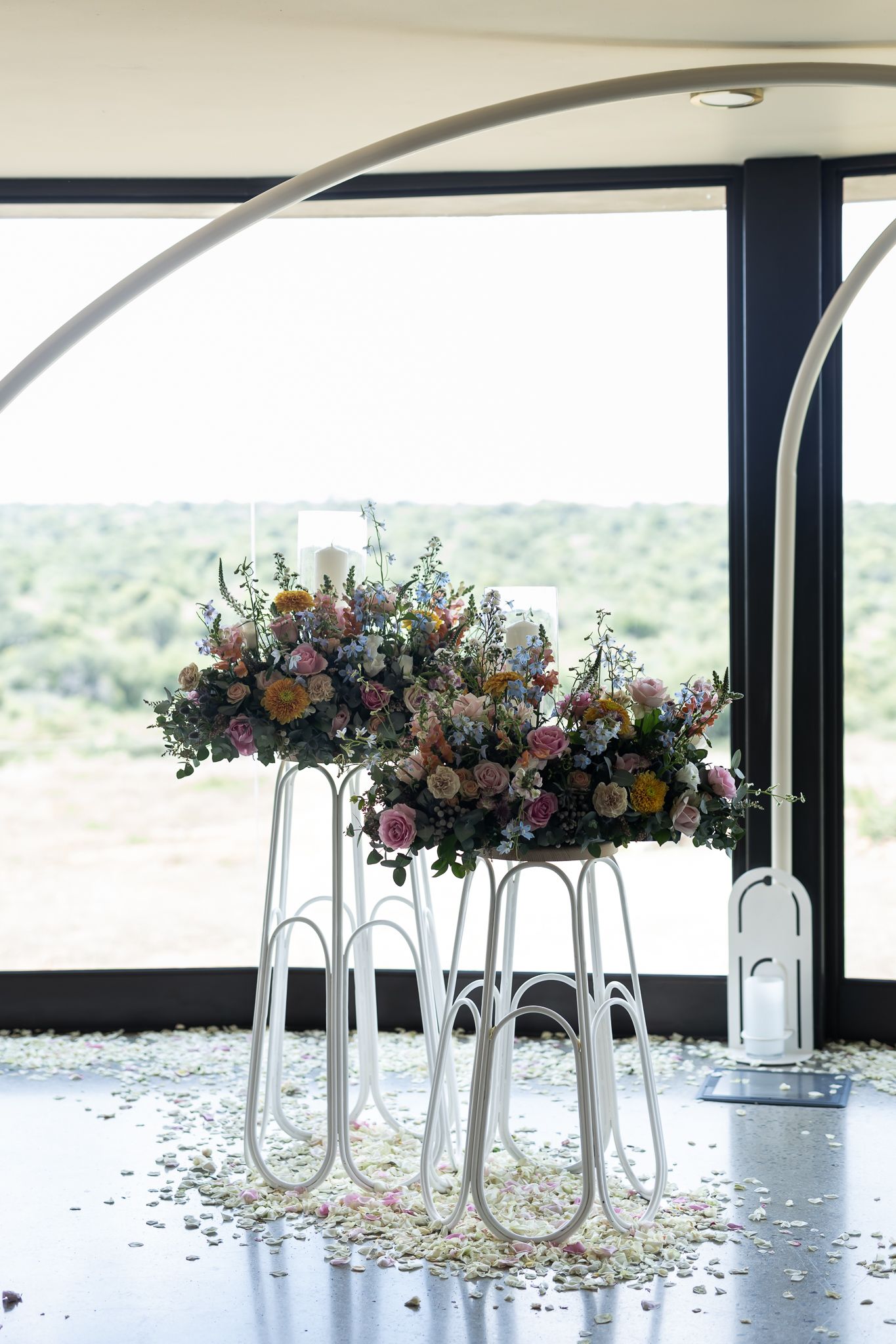 Modern floral arrangements on white stands with window view of landscape in background.