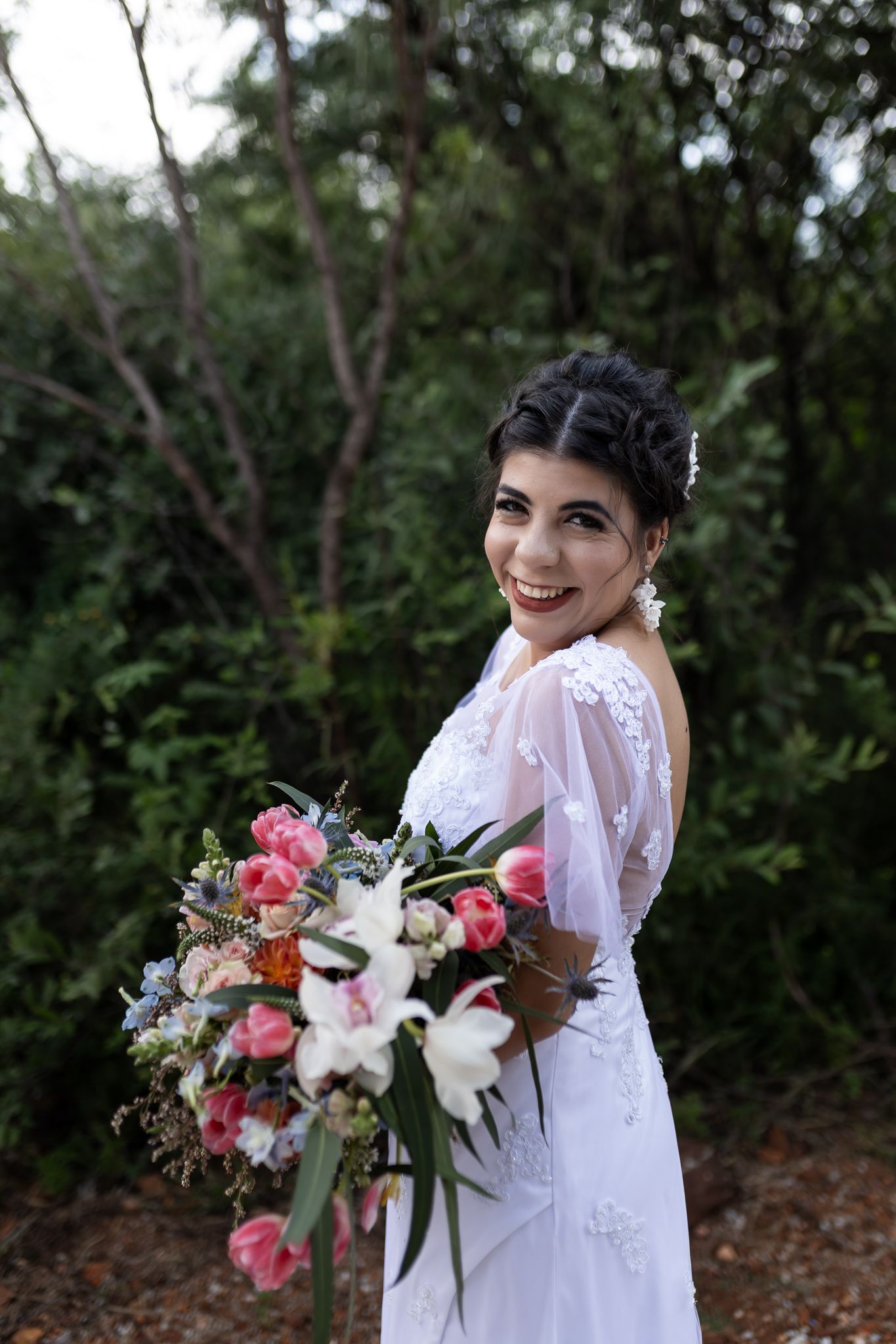 A bride in a white lace gown poses with her bouquet against a natural outdoor background.