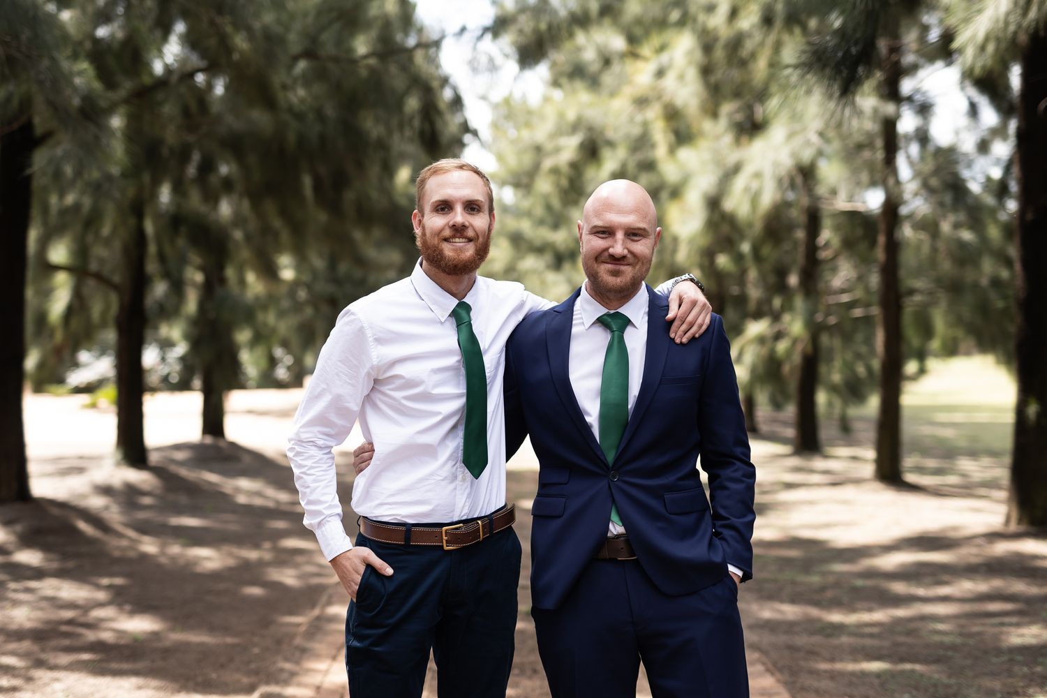 Two groomsmen in white shirt and navy suit share friendly moment in pine forest.