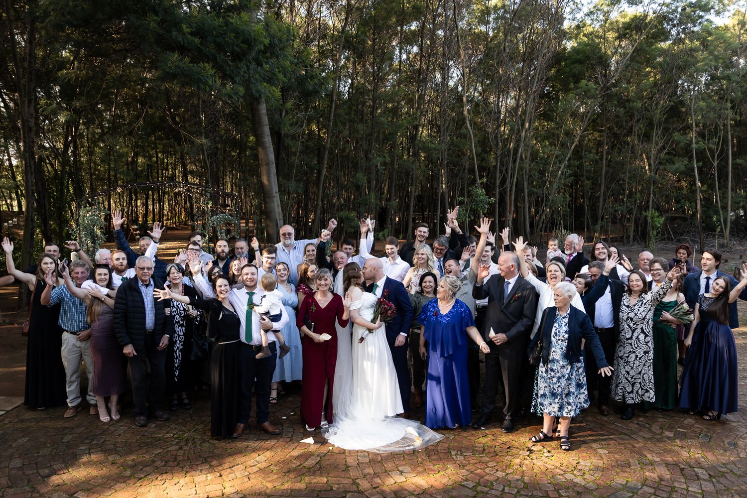 Large group wedding photo with guests gathered in a forest clearing.