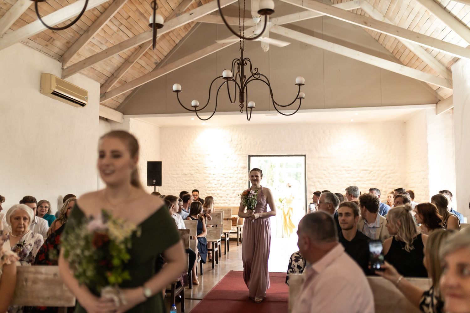 Wedding ceremony in progress inside a bright chapel with elegant chandeliers and wooden beam ceiling.