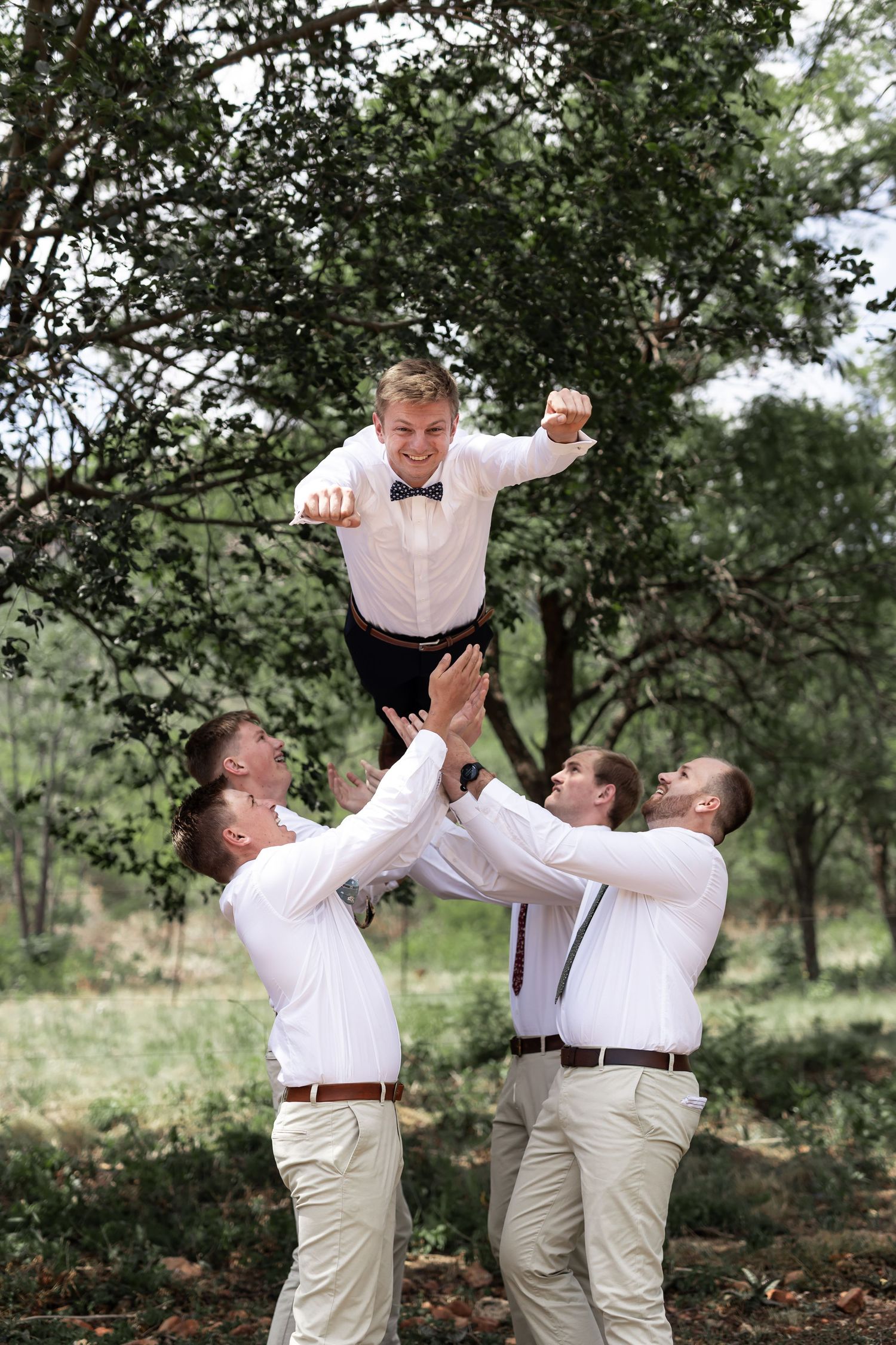 Wedding party members lift and toss groomsman in playful outdoor photo.