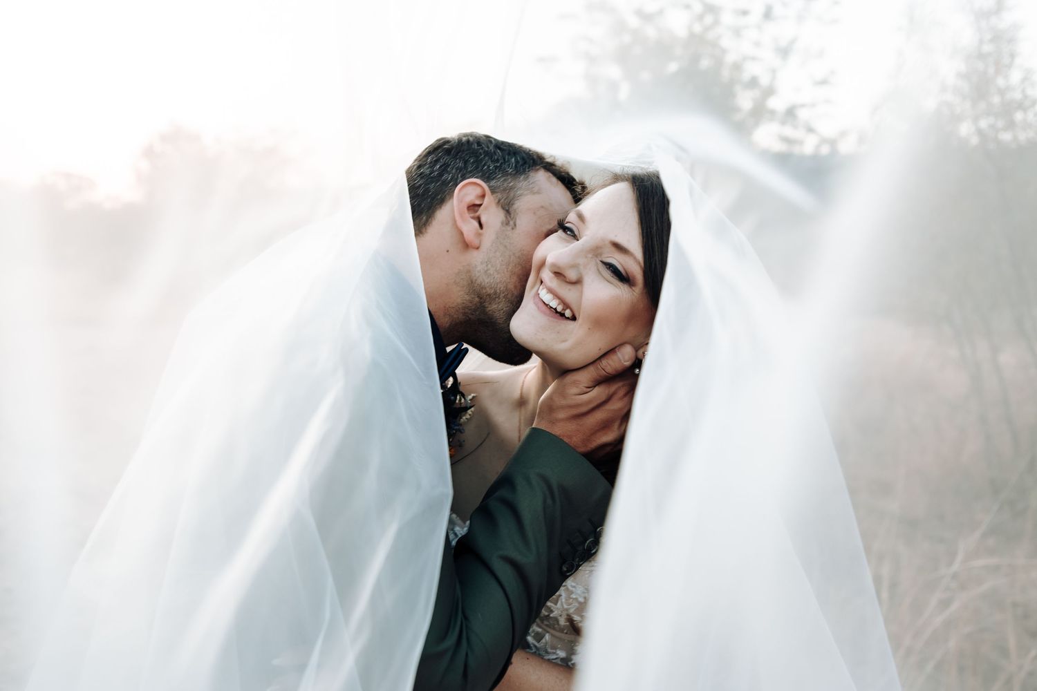 Joyful couple shares a laugh under bridal veil in soft sunlight.